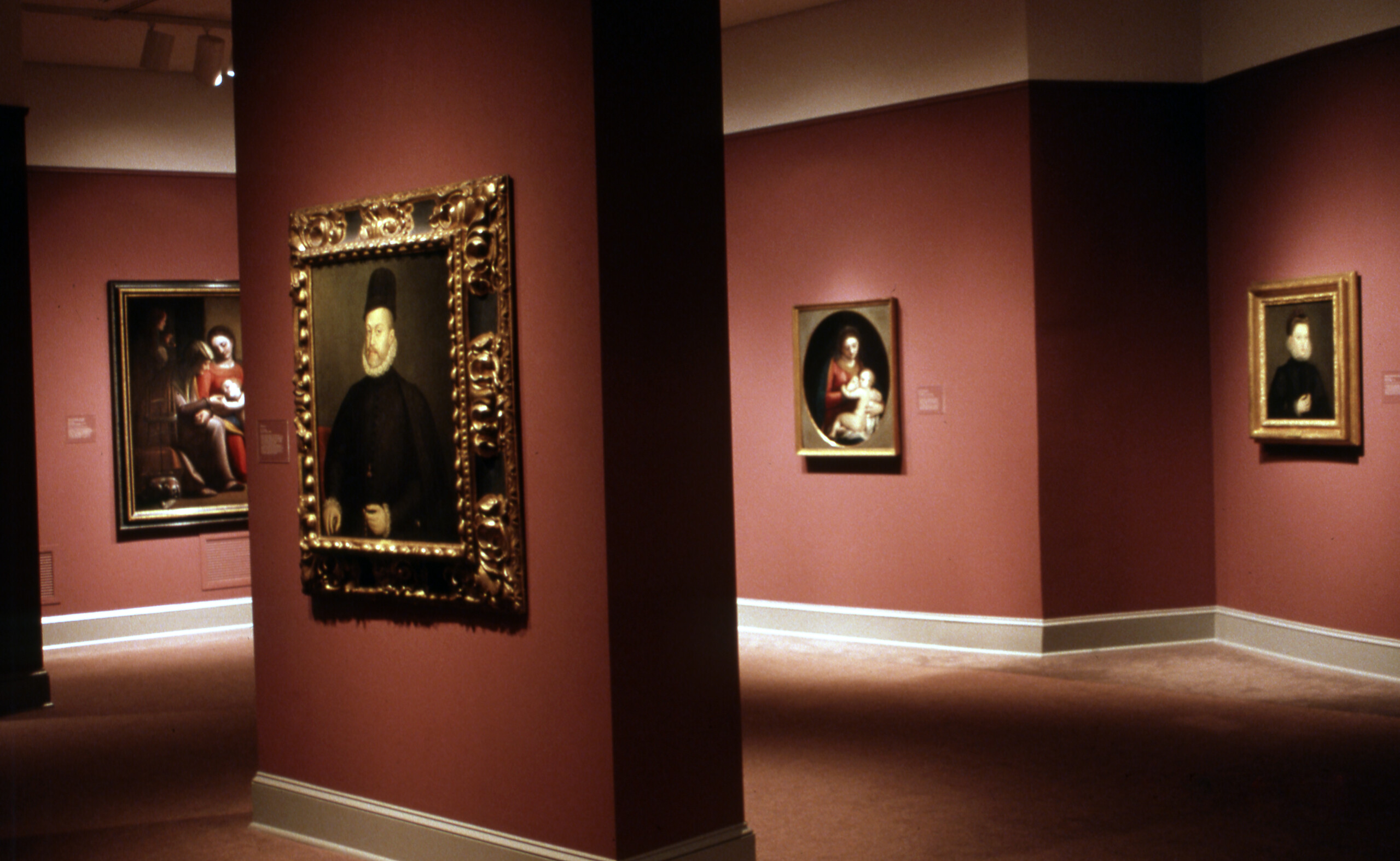 A view of a gallery space with dark red walls. On each wall, a painting is hanging. The paintings are portraits as well as biblical scenes.