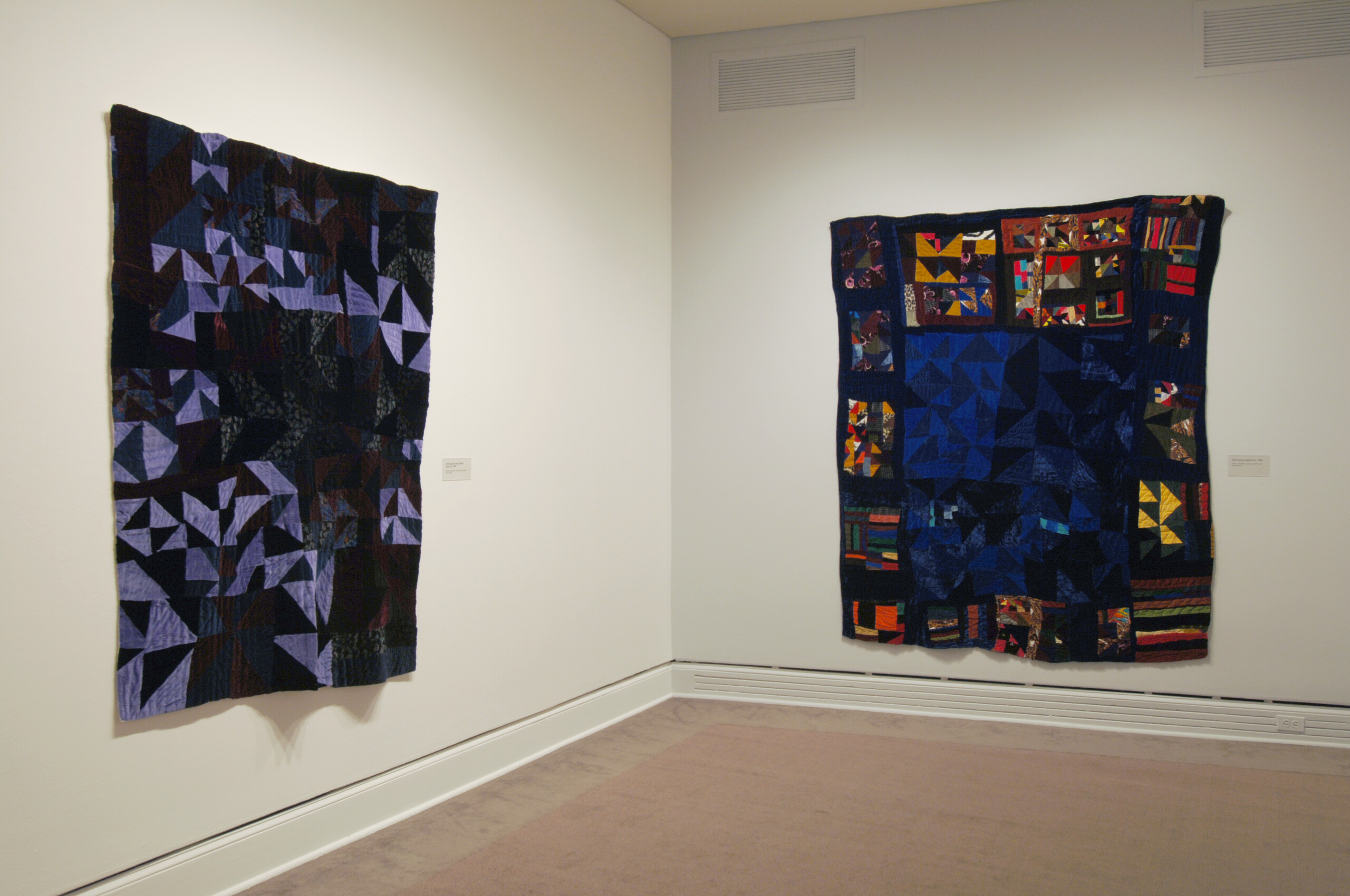 An installation view of a gallery space shows two large quilts hanging on a white wall. The quilts have an abstract pattern and have dark colors, especially the dark blue colors are prominent, creating a feeling of nighttime.