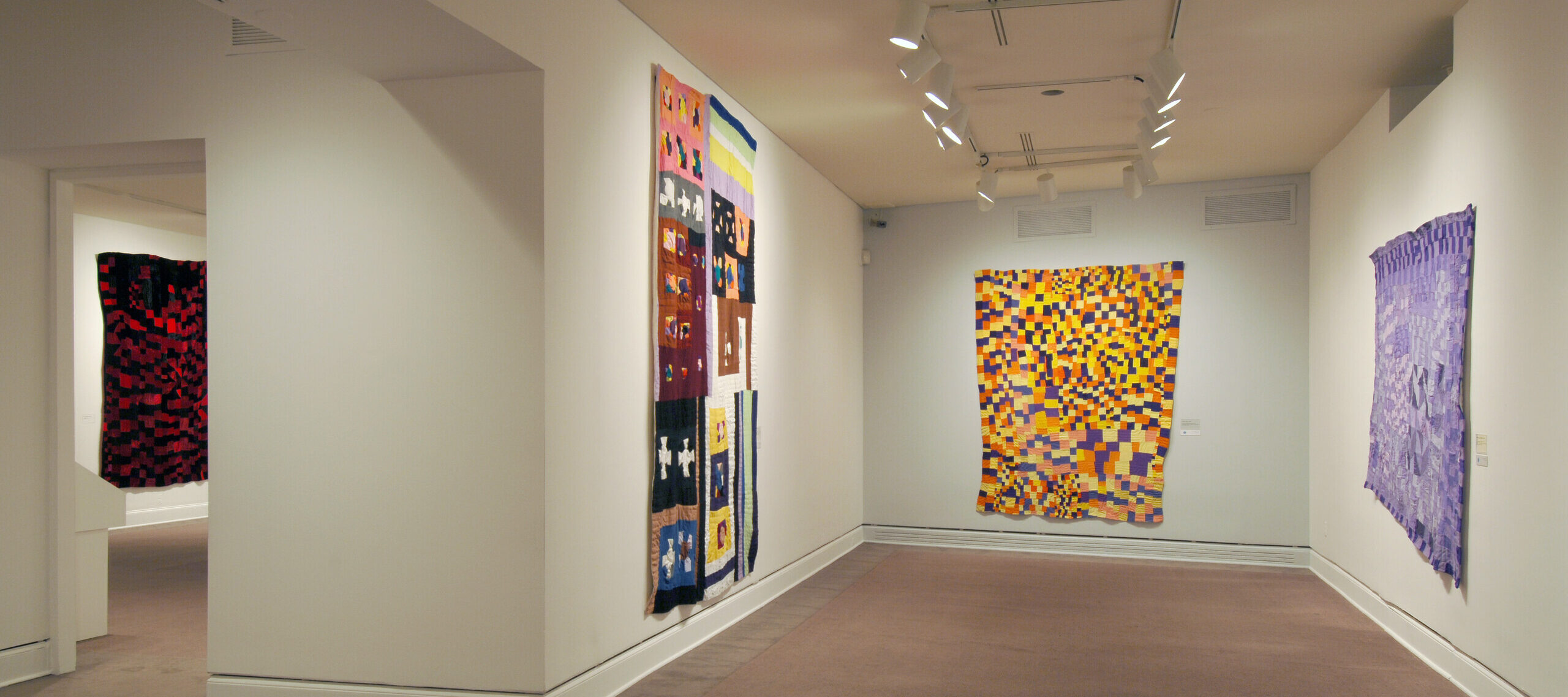 A view of a gallery space. Large quilts are hanging on each wall. They are colorful and almost take up the whole wall, creating a warm and cozy interior.