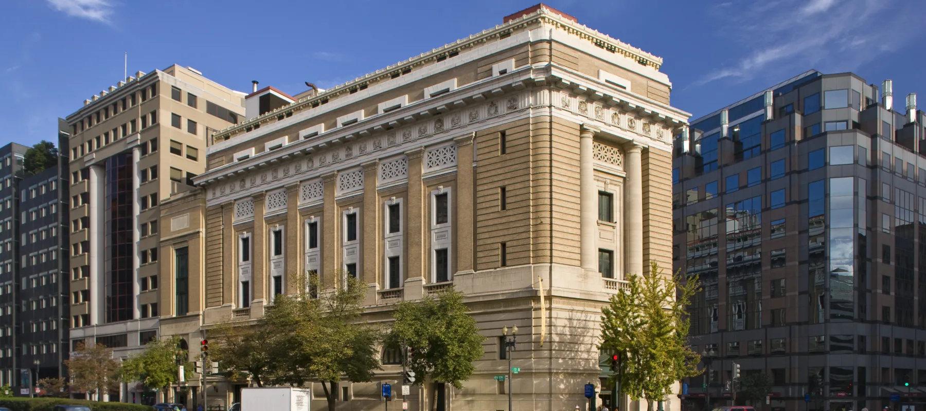 View of the museum from outside showing the Neoclassical building from one corner. The building is a tan-colored stone with an arched doorway, long vertical windows, and detailed molding around the roof.