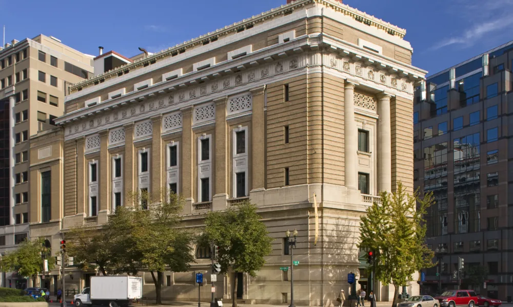 View of the museum from outside showing the Neoclassical building from one corner. The building is a tan-colored stone with an arched doorway, long vertical windows, and detailed molding around the roof.