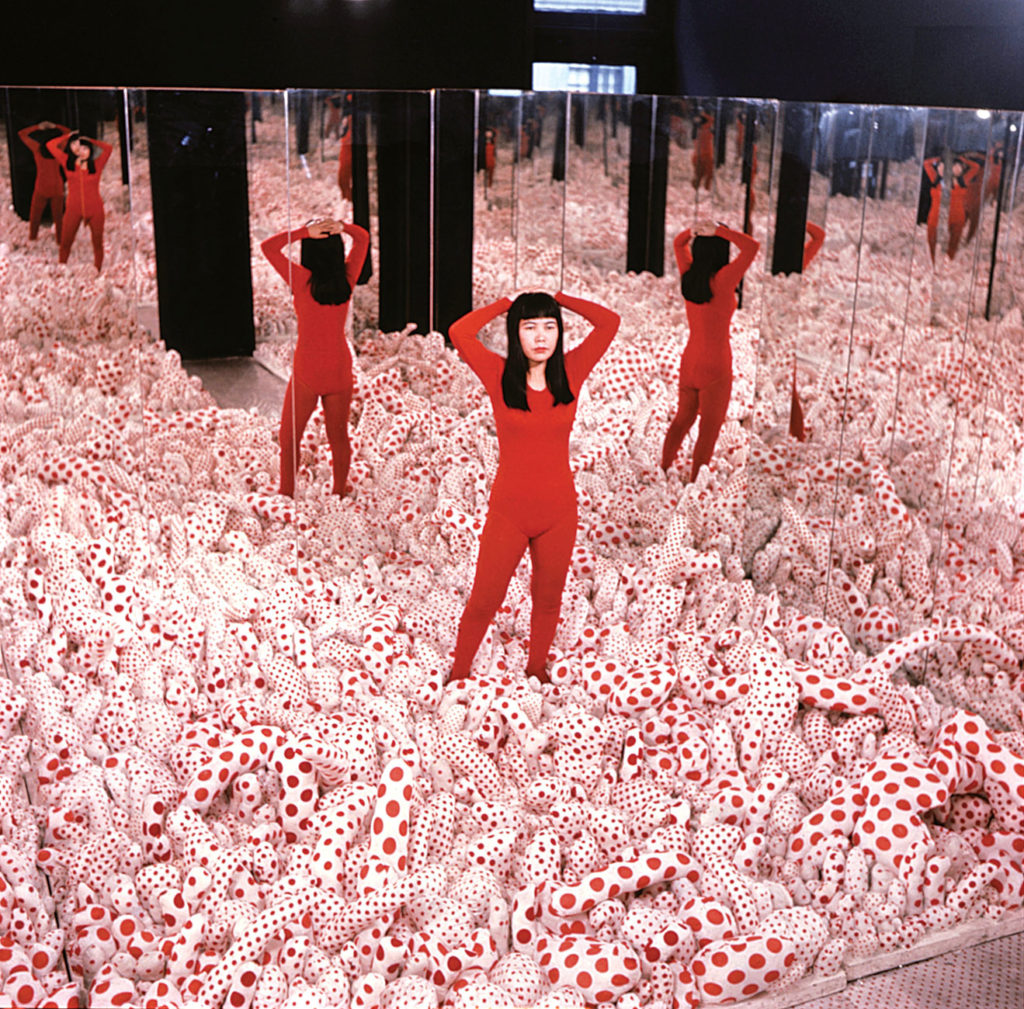 A woman with a light skin tone and long black hair is standing in a room with mirrors on the wall and thousands of white objects that are covered in red polka dots lying on the floor. She wears a red body suit that connects to the red of the polka dots and holds her arms behind her hear.