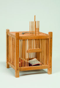 A square cage made out of light-colored wood with a wire handle on top. The opening gate is slid open to reveal a book flipped open within the cage.