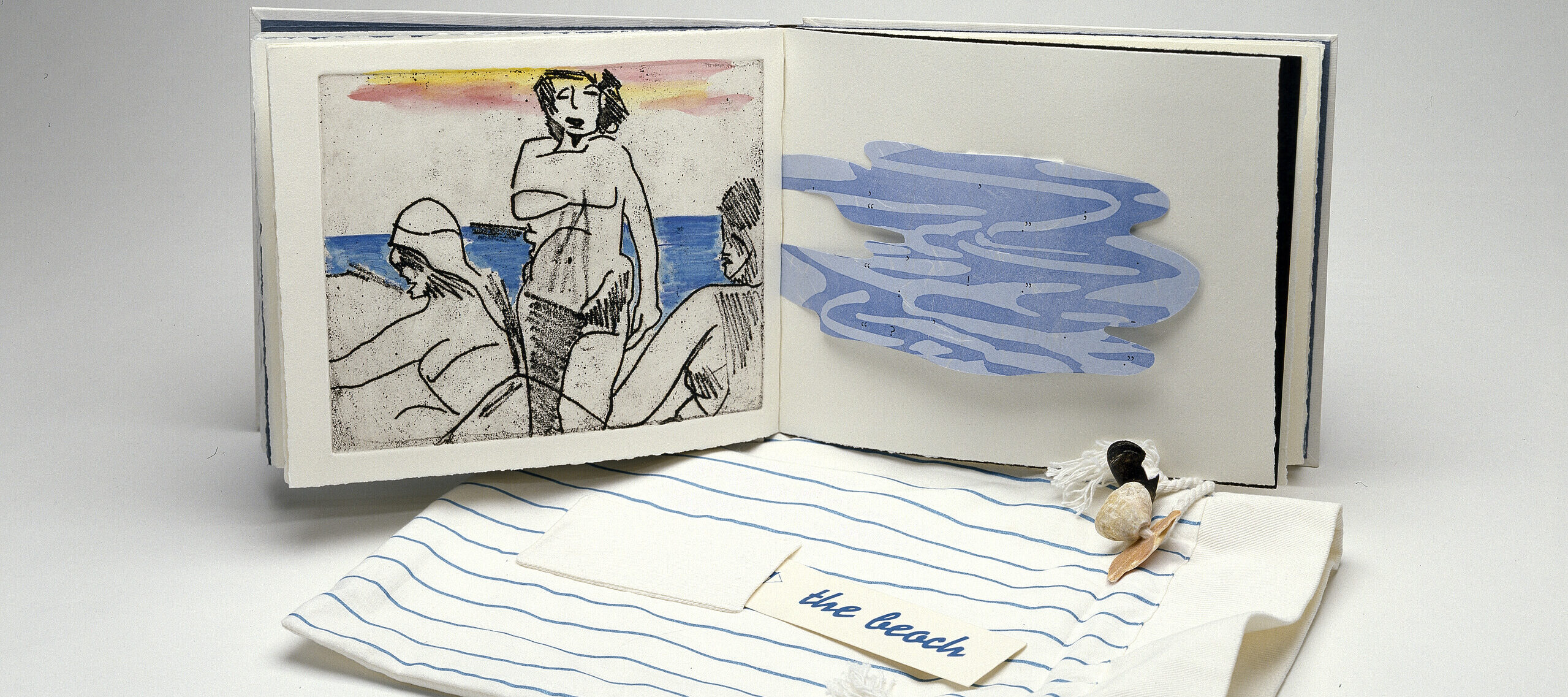 An open book is in the background and its cover is in the foreground. The left book page features a print of three figures on a beach and the right book page has a blue, amorphous shape with wavy lines. The book cover is a drawstring bag made of striped blue and white fabric. On top, it has a pocket that holds a card which reads “the beach” and is ornamented with seashells.