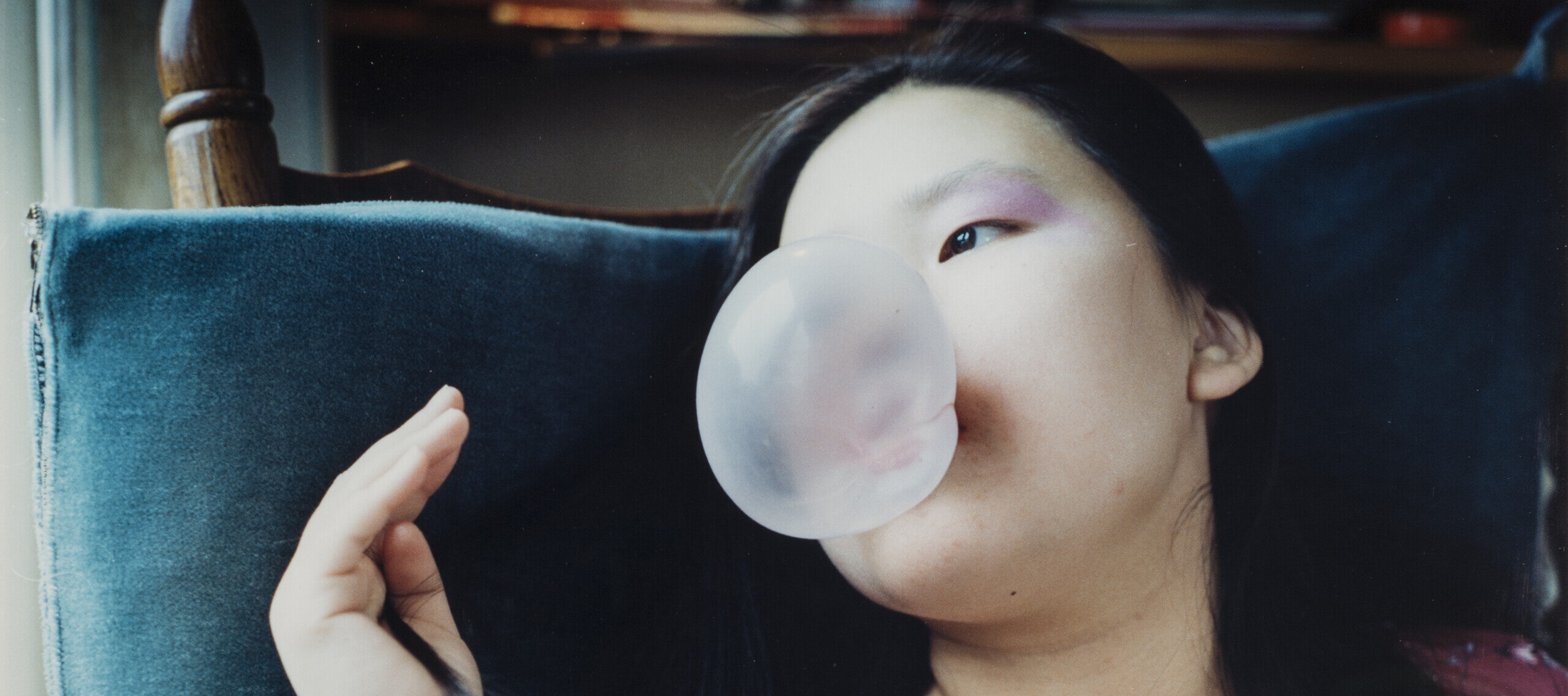A color photograph of a light skinned person wearing a sheeer floral dress sitting on a chair and reclining back on a large blue pillow. They are blowing a large bubblegum bubble while gazing to the left of the frame.