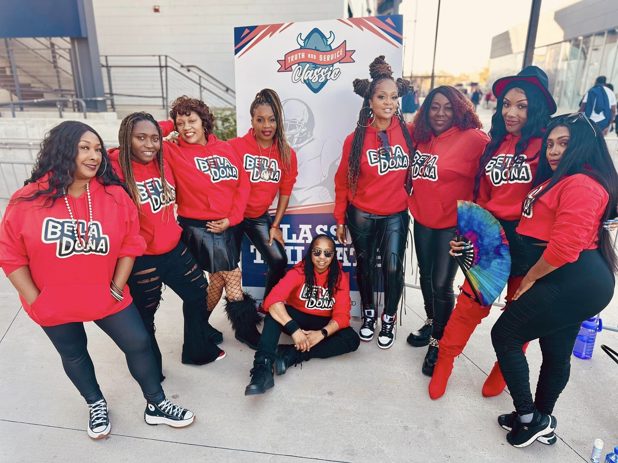 Nine medium-skinned women pose together wearing matching red sweatshirts with "Be'la Dona" printed on them in black and white letters.