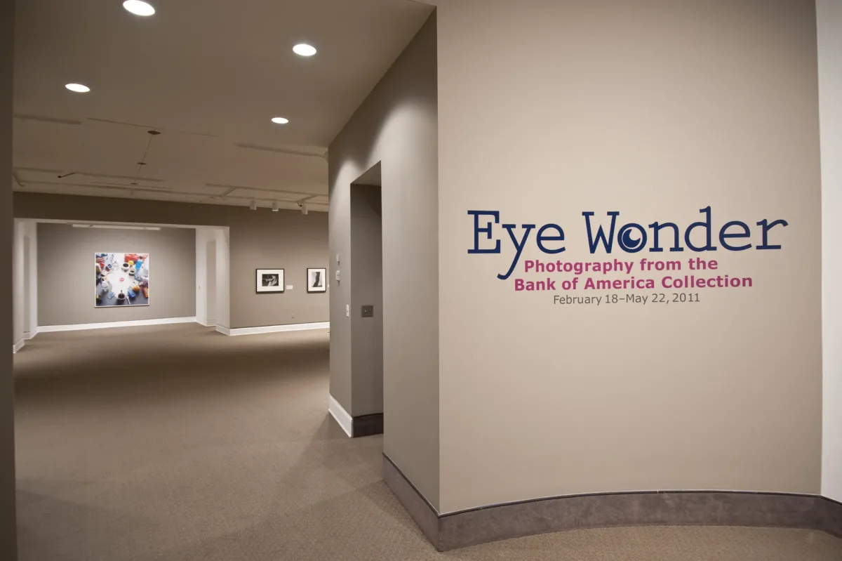 An installation view of a gallery space with white walls and a gray floor. On the wall facing the viewer it says "Eye Wonder: Photography from the Bank of America Collection" in blue and pink letters.