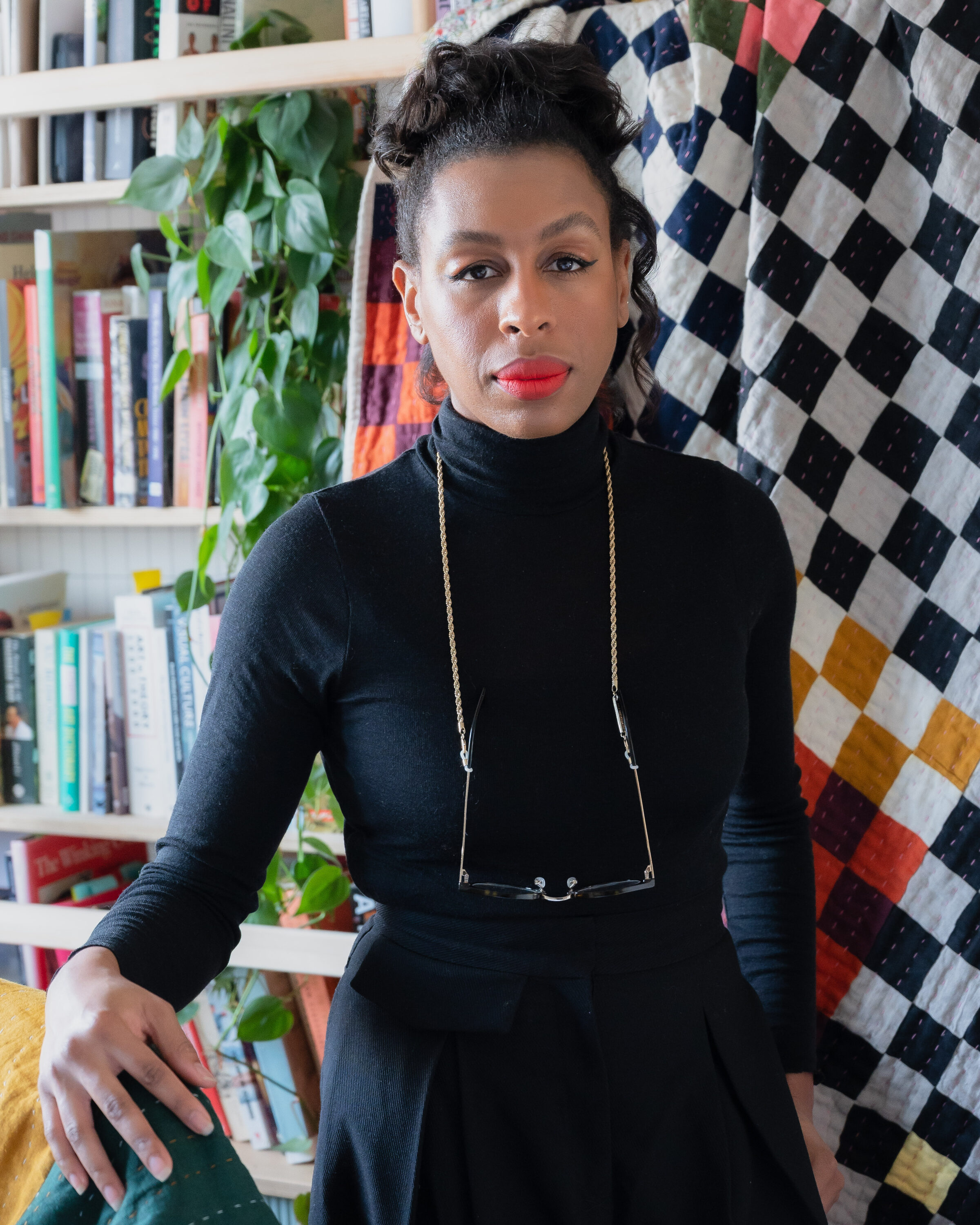 A dark-skinned woman wearing a black turtle neck and skirt stands in front of a colorful checkered quilt, which hangs from a white bookcase. A lush plant is situated among the books. The woman stares at the camera. Her hair is worn atop her head, she has on red liptstick, and wears her glasses around her neck on a thin chain.