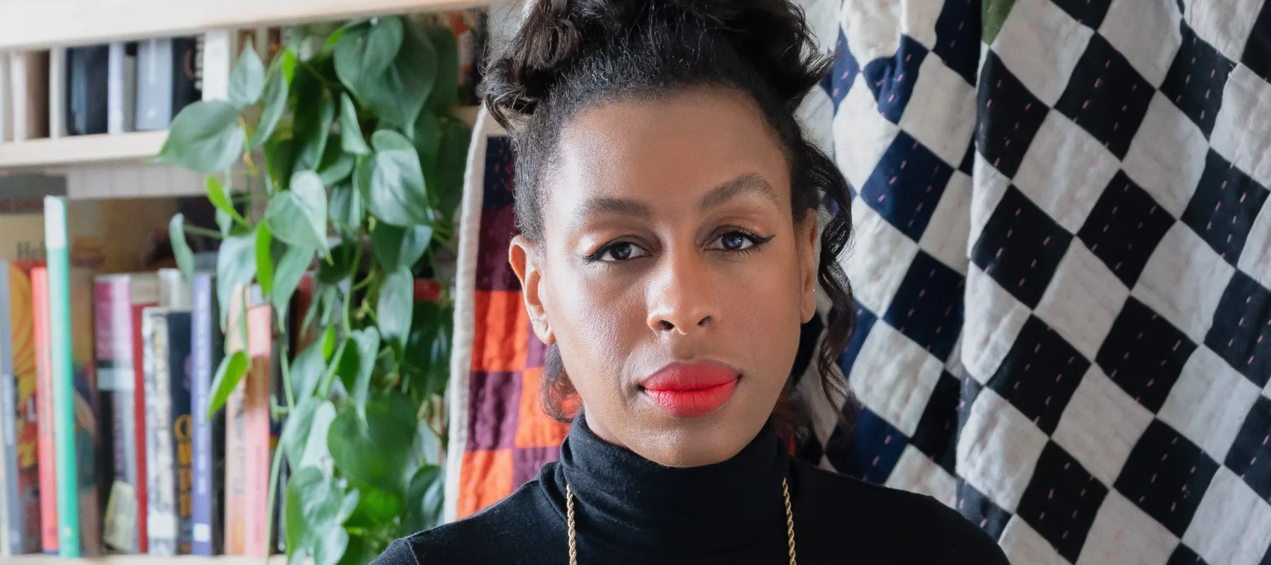 A dark-skinned woman wearing a black turtle neck and skirt stands in front of a colorful checkered quilt, which hangs from a white bookcase. A lush plant is situated among the books. The woman stares at the camera. Her hair is worn atop her head, she has on red liptstick, and wears her glasses around her neck on a thin chain.