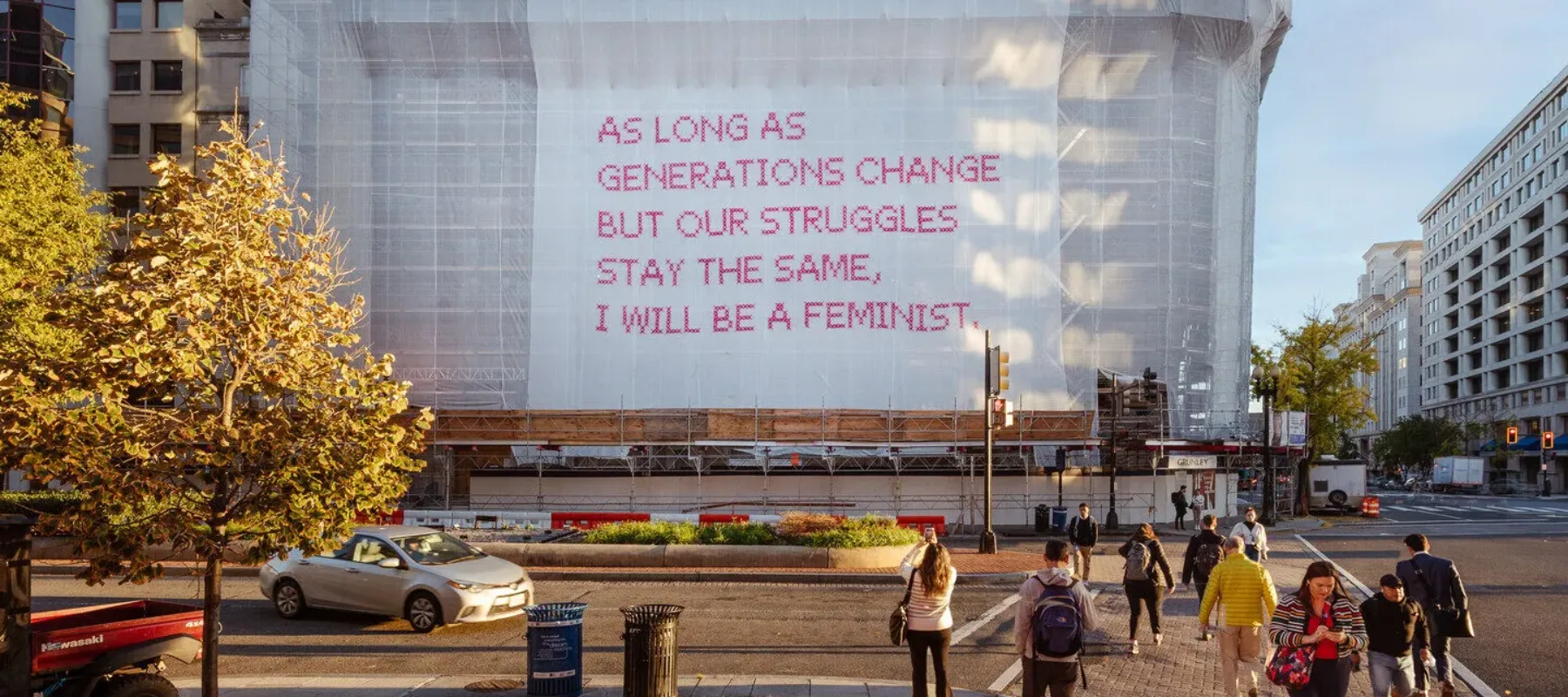 A building with a white mesh artwork covering its façade, featuring bright pink cross-stitched letters that say "As long as generations change but our struggles stay the same, I will be a feminist."