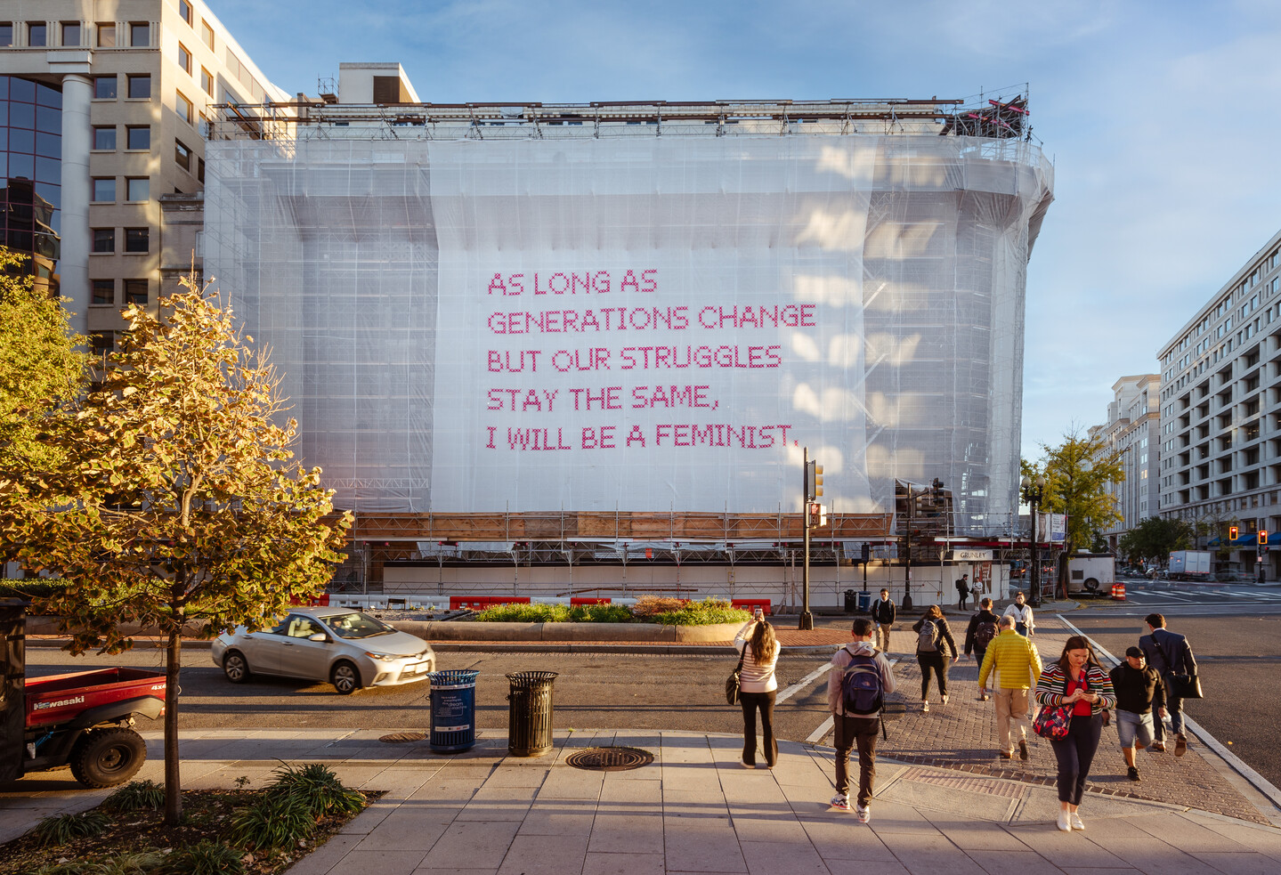 A building with a white mesh artwork covering its façade, featuring bright pink cross-stitched letters that say "As long as generations change but our struggles stay the same, I will be a feminist."