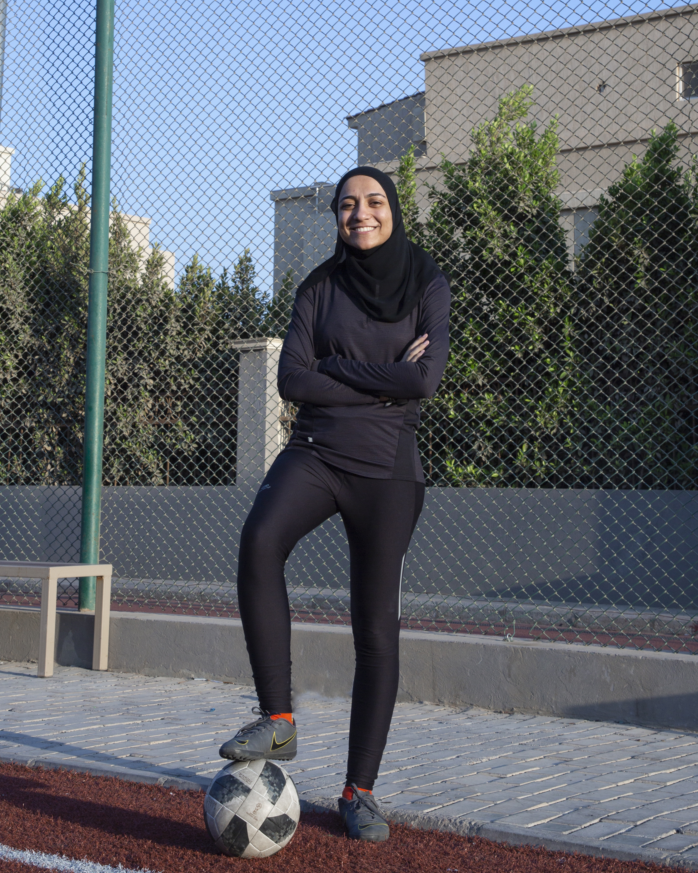A woman stands outdoors, dressed in black workout gear and head covering. She smiles at the viewer with her arms crossed and right foot resting on a soccer ball.