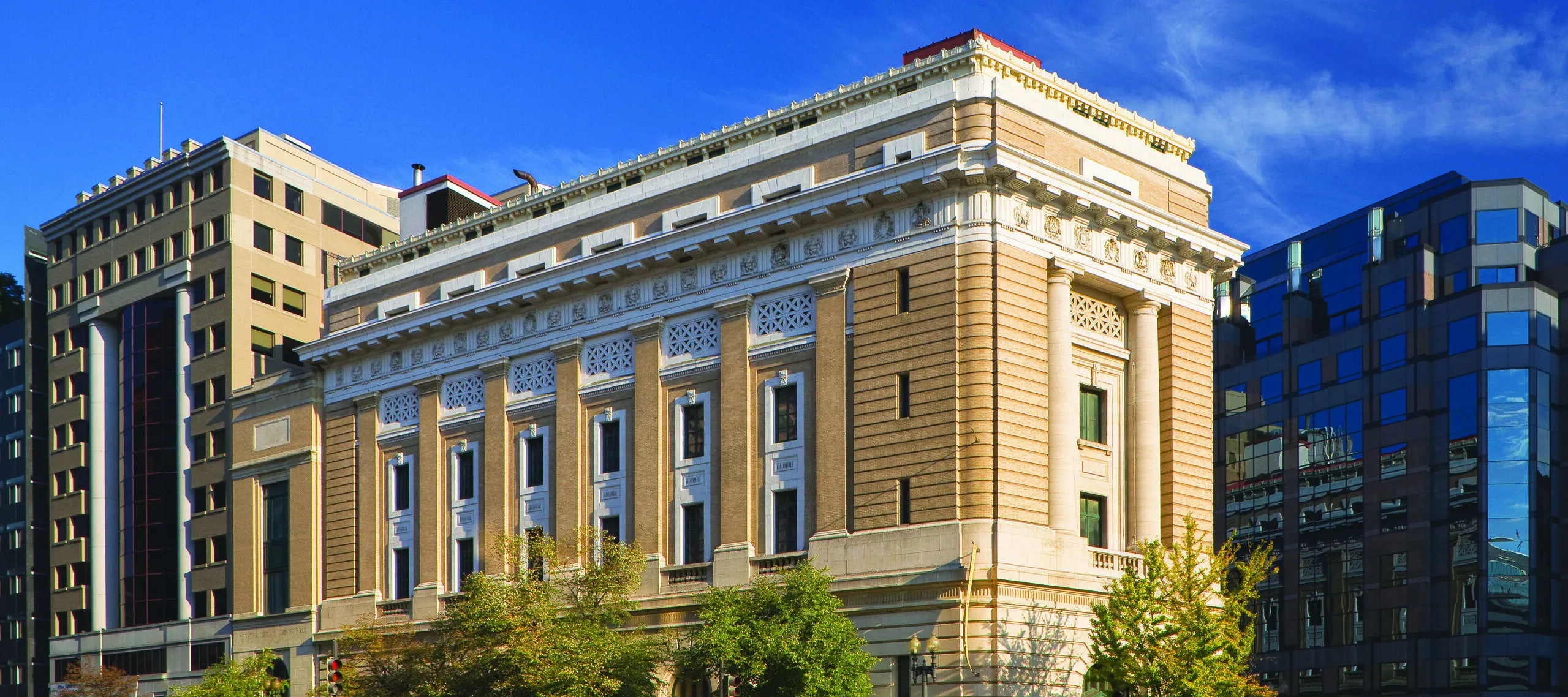 View of the National Museum of Women in the Arts building from outside showing the Neoclassical building from one corner. The building is a tan-colored stone with an arched doorway, long vertical windows, and detailed molding around the roof.