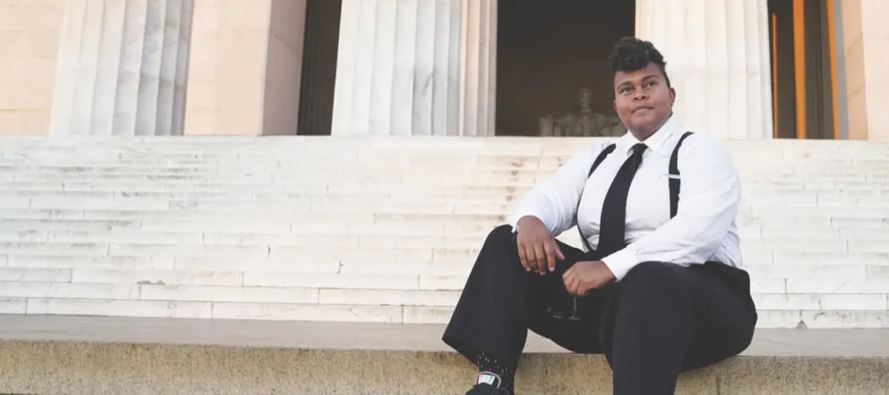 A person with medium skin tone sits on the stairs of the Lincoln Memorial, a white stone building with fluted columns.