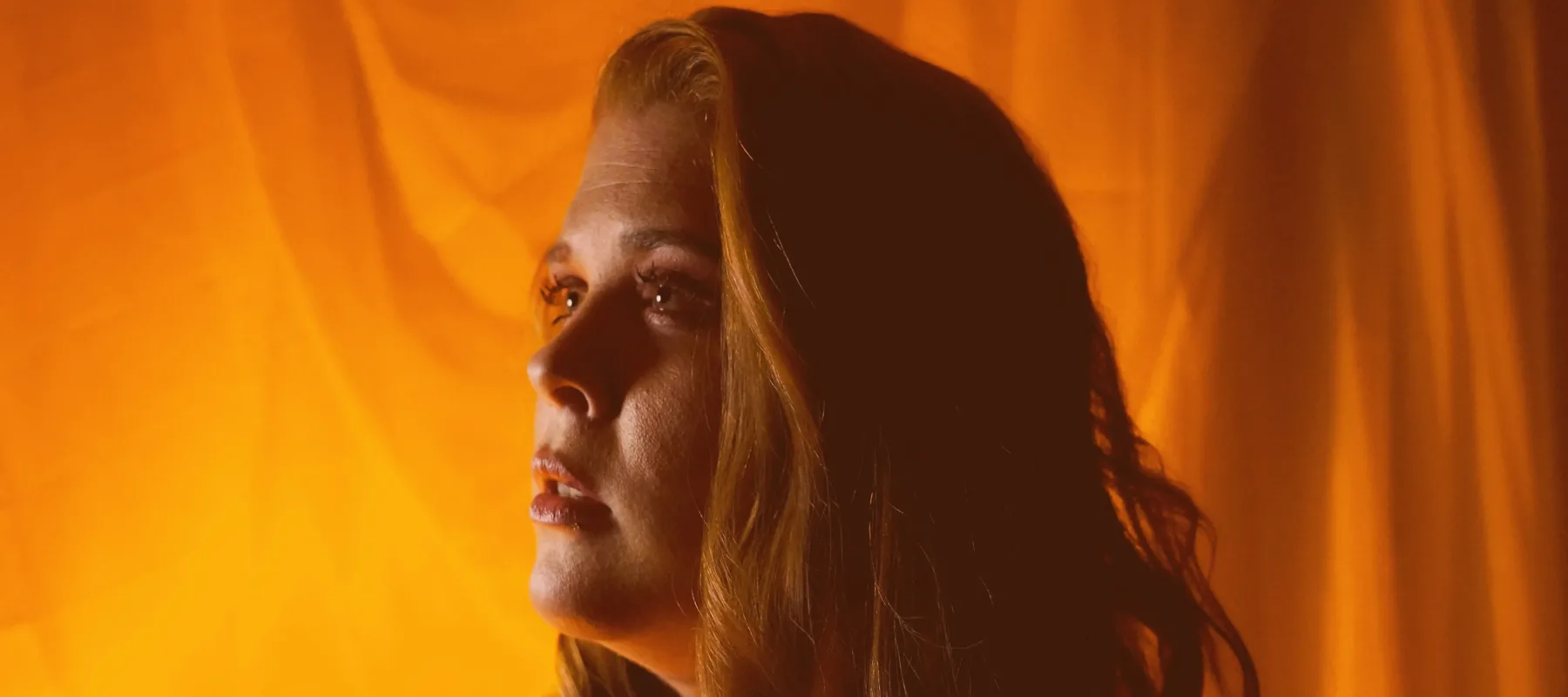 A light-skinned person with red hair looks off camera. The half of their face closest to the camera is in shadow, with light coming across the other side of their face. Behind them is an orange background.