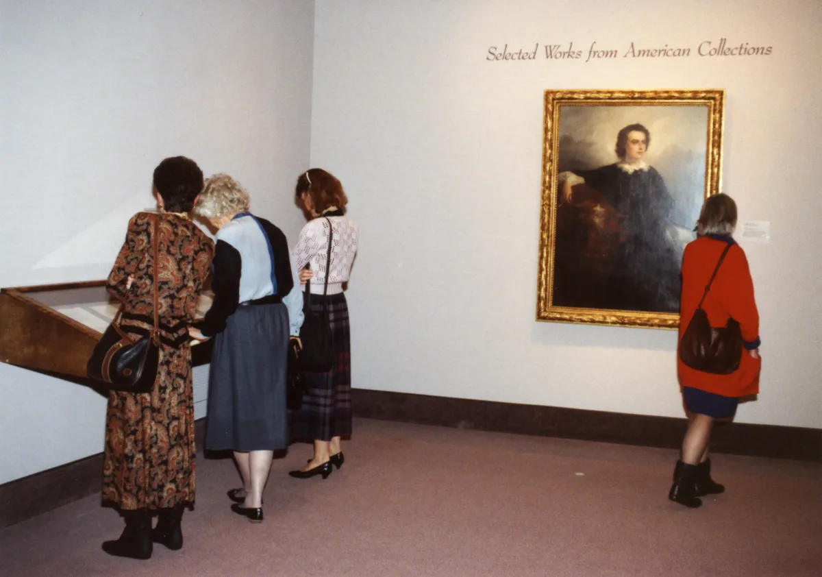 View of a gallery space. Several people are in the museum. On one wall, a portrait is hanging underneath the text "Selected works from the American Collections."