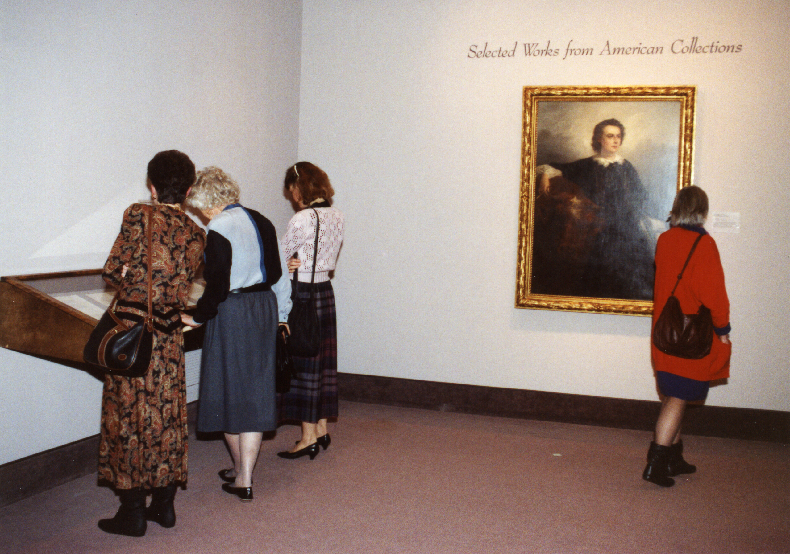 View of a gallery space. Several people are in the museum. On one wall, a portrait is hanging underneath the text 