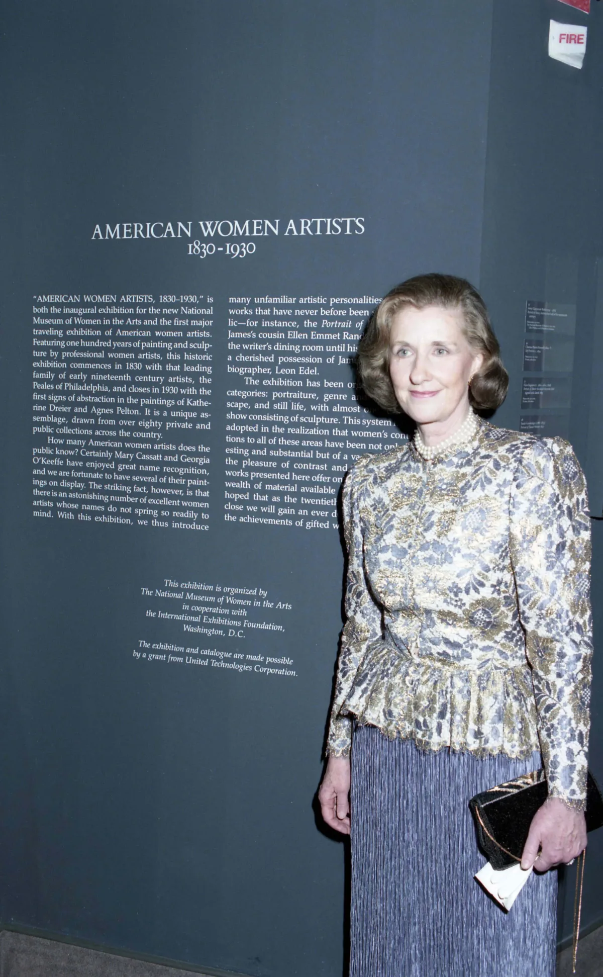 A woman with a light skin tone and grayish-blonde hair is standing next to a blue wall. She is wearing a floral blouse, a long skirt, and pearls, smiling at the camera. On the wall, it says in big, white letters "American Women Artists: 1830-1930."