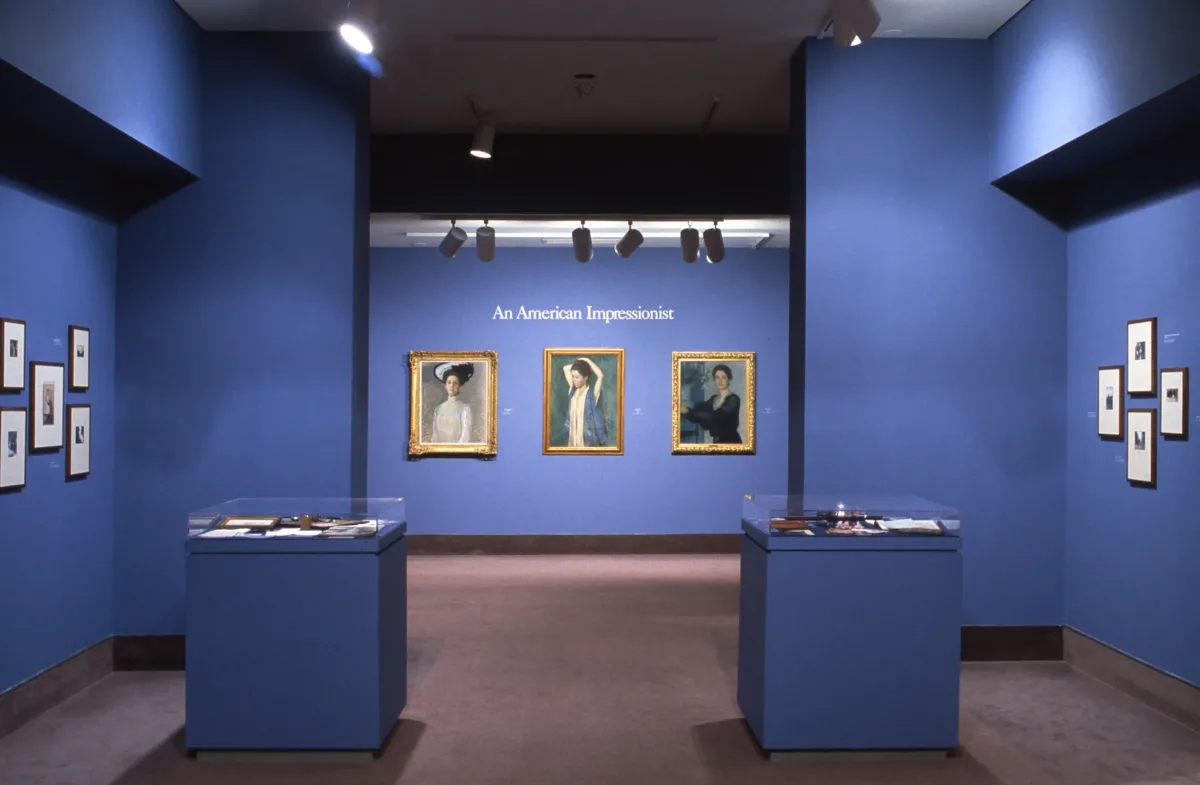 A view of a gallery shows several oil paintings hanging on blue walls. Three portraits of women in an Impressionist painting style hang next to each other. In large letters, it says "An American Impressionist" above the paintings.