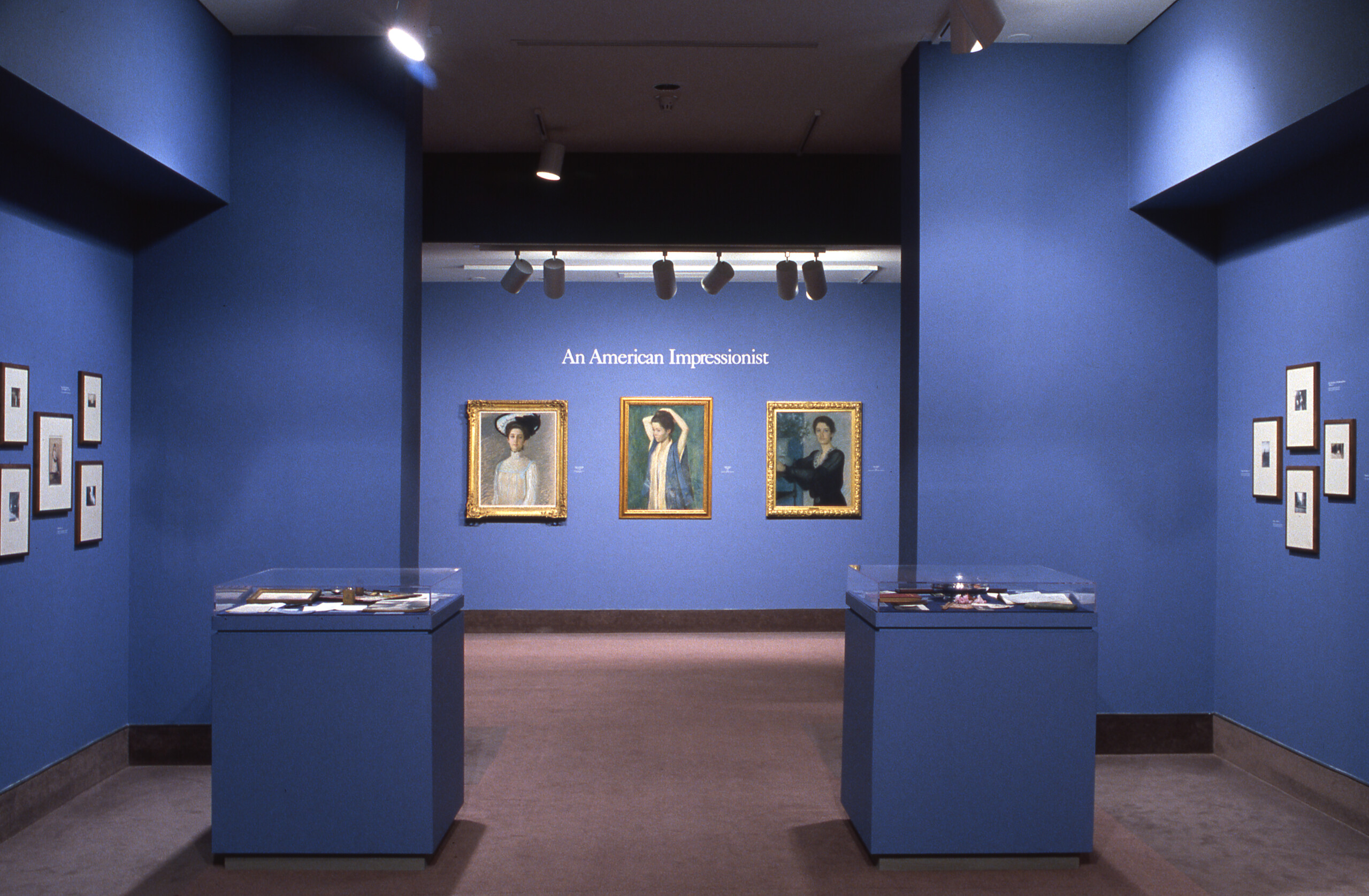 A view of a gallery shows several oil paintings hanging on blue walls. Three portraits of women in an Impressionist painting style hang next to each other. In large letters, it says 