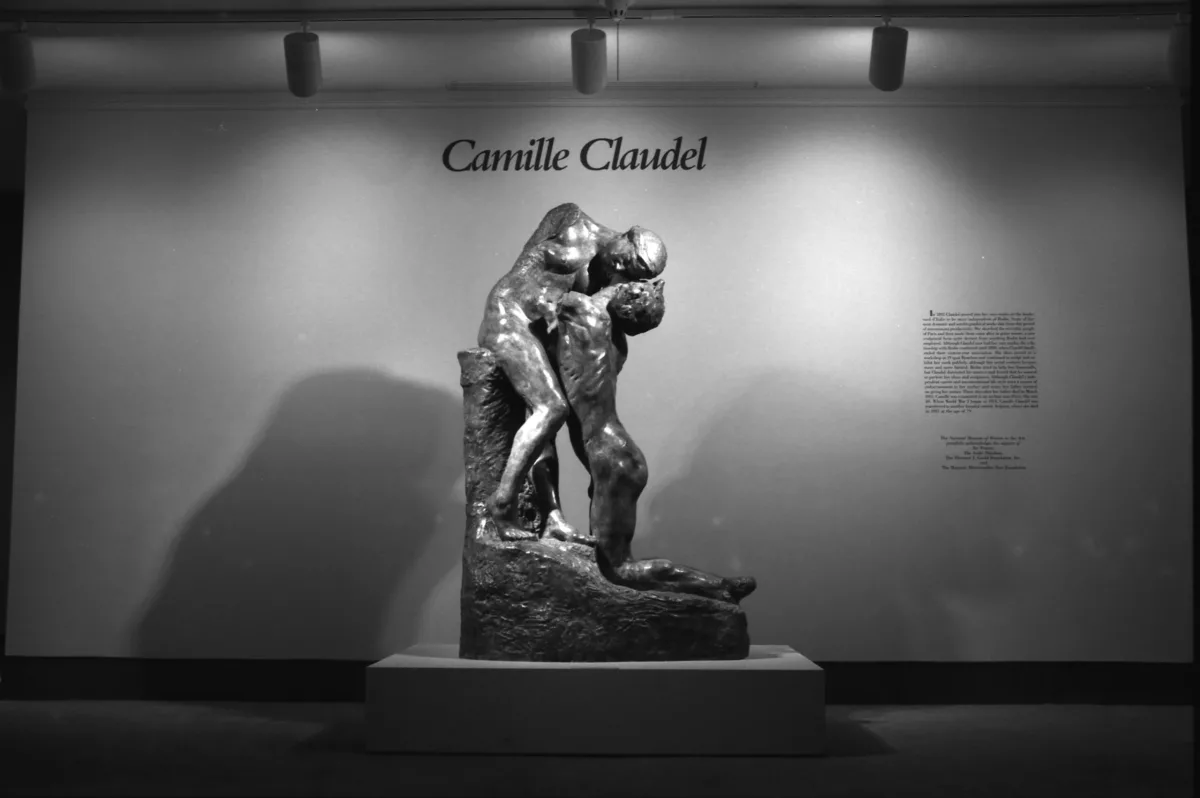 A view of a gallery space. Two sculptures of a man and a woman are placed on a pedestal. The woman is leaning down to the man, who is on his knees, and kissing him. Above the sculptures, it says "Camille Claudel."