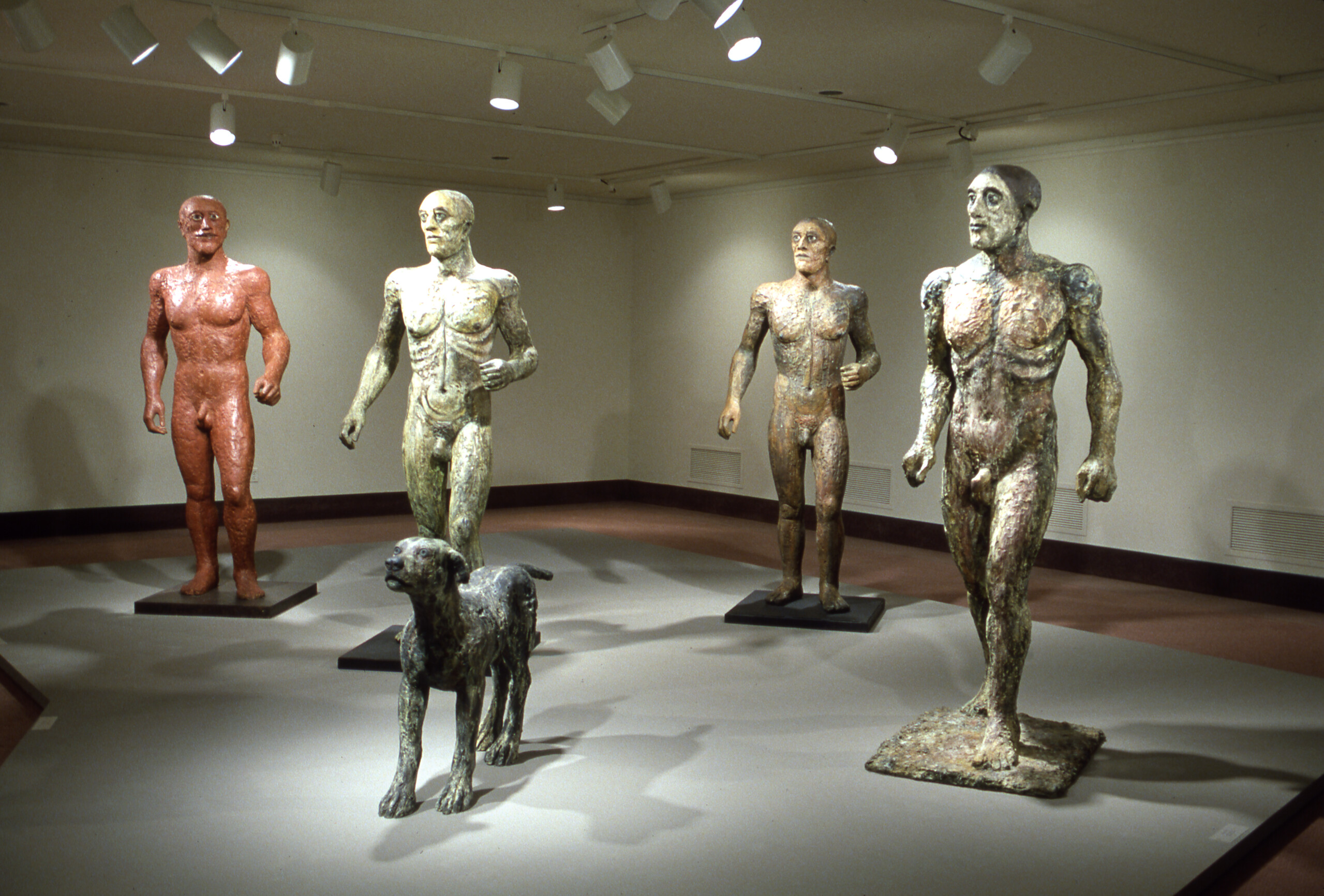 A view of a gallery space. Four tall sculptures of men seem to be running through the gallery. A dog is walking in front of them.