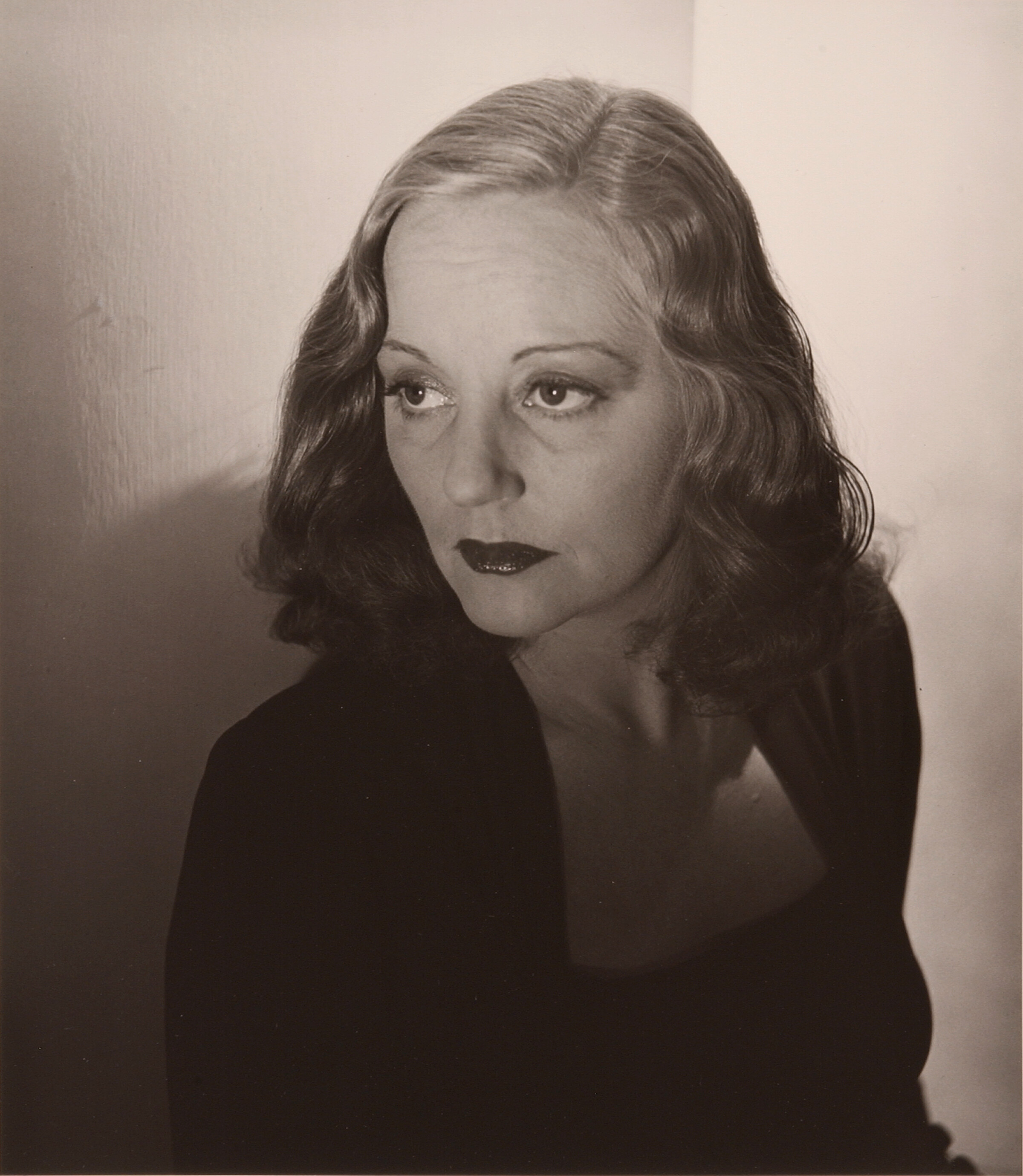 A black-and-white photograph of a woman staring coldly off frame, to the left. She has thin eyebrows and wears dark lipstick; her light hair is worn at shoulder-length, with subtle waves. She wears a dark cardigan over a dark, low-cut top. The photograph’s lighting is dramatic, with shadow in the bottom half, and light from the woman’s eyes upward. 
