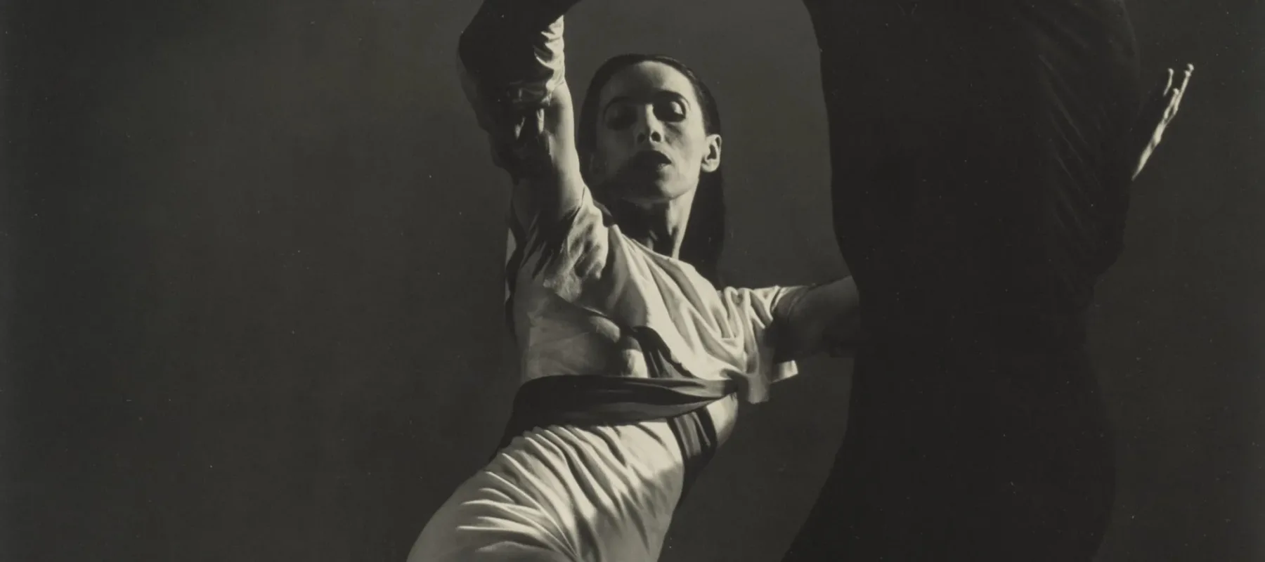 Black-and-white photograph of a man, dressed in black, and Martha Graham, in white, dancing. Graham, lit from above, strikes one of her famous angular poses, both legs bent, the back foot pointing behind her. Her raised arms are held by the man, who stand