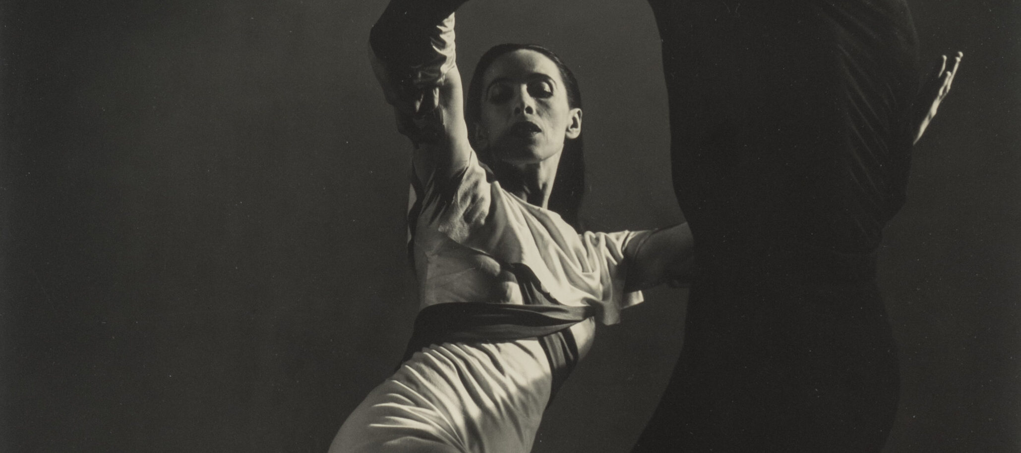 Black-and-white photograph of a man, dressed in black, and Martha Graham, in white, dancing. Graham, lit from above, strikes one of her famous angular poses, both legs bent, the back foot pointing behind her. Her raised arms are held by the man, who stand