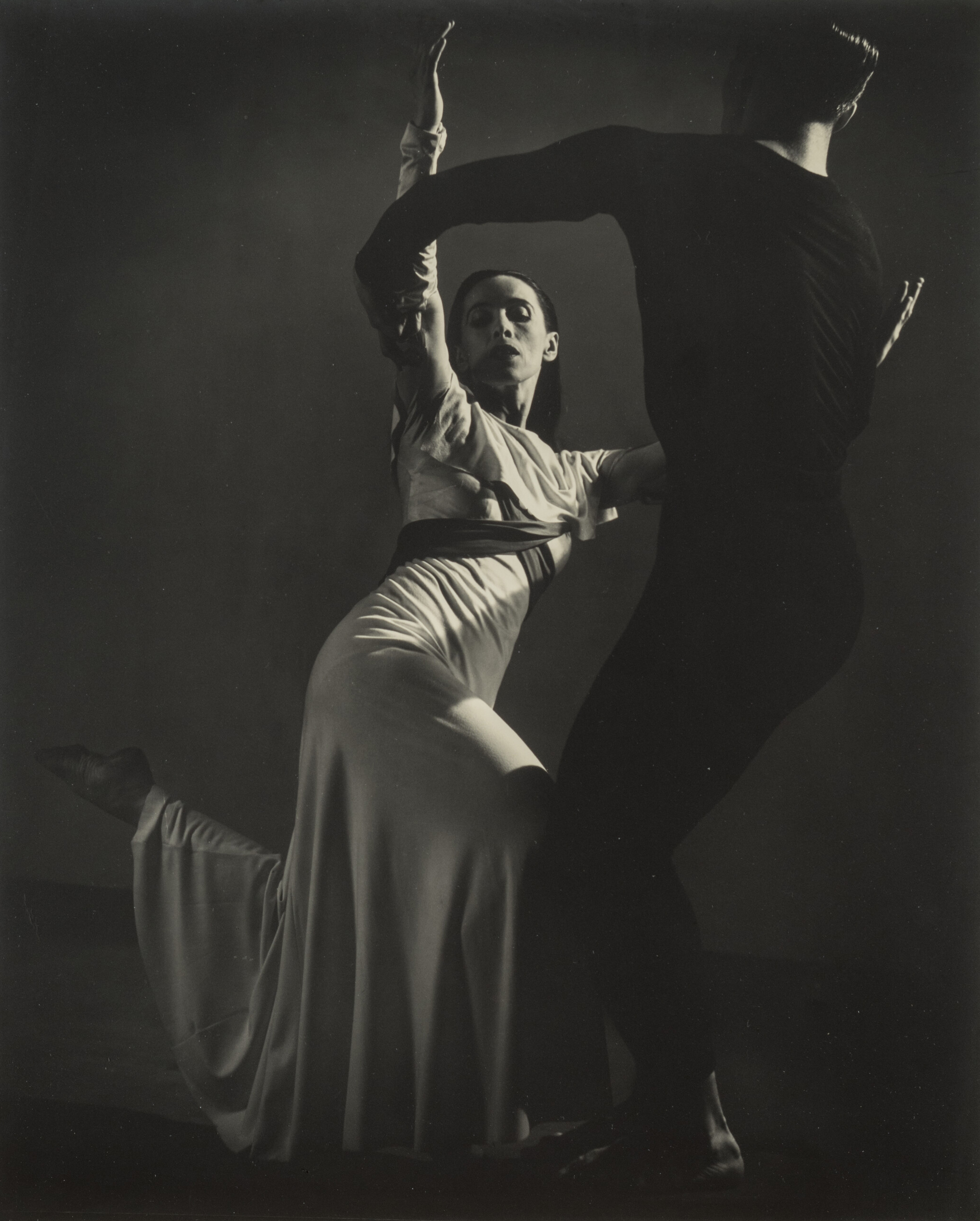 Black-and-white photograph of a man, dressed in black, and Martha Graham, in white, dancing. Graham, lit from above, strikes one of her famous angular poses, both legs bent, the back foot pointing behind her. Her raised arms are held by the man, who stand