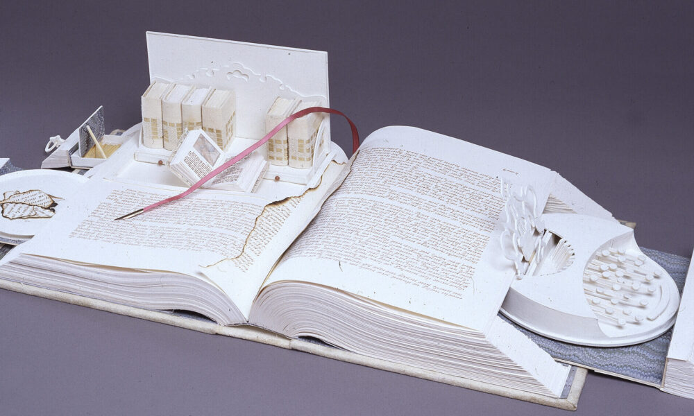 A white book opens like a Swiss army knife; its pages cut to create a sculpture. Opened to the center, pull-out drawers open from the sides, filled with paper-made objects. On the right, a typewriter-like object. On the left, a box with smaller books, including one that is also open.