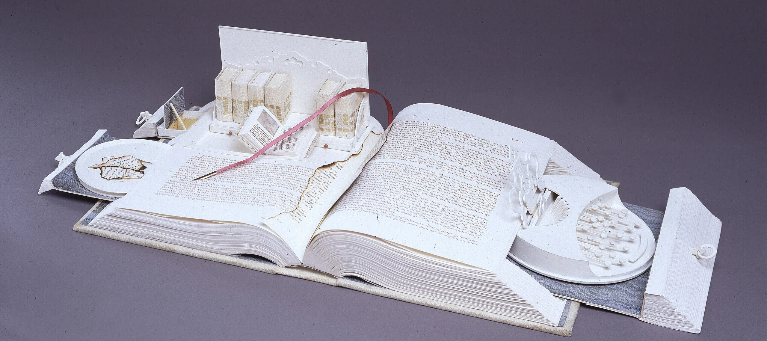 A white book opens like a Swiss army knife; its pages cut to create a sculpture. Opened to the center, pull-out drawers open from the sides, filled with paper-made objects. On the right, a typewriter-like object. On the left, a box with smaller books, including one that is also open.