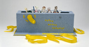 Paper figures in swimwear emerge from a grey-blue box with a slot in the top, like a toaster, and a yellow crank on the side. Small signs say “talking” “and laughing” “exercise” “class.” Yellow ribbons, untied, are at the base of the box.