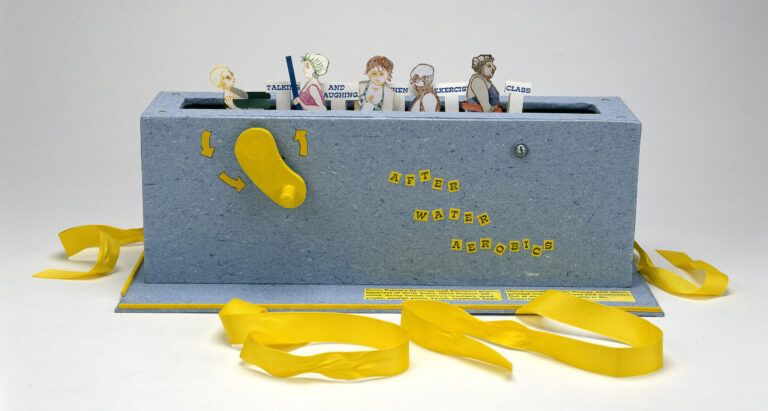 Paper figures in swimwear emerge from a grey-blue box with a slot in the top, like a toaster, and a yellow crank on the side. Small signs say “talking” “and laughing” “exercise” “class.” Yellow ribbons, untied, are at the base of the box.