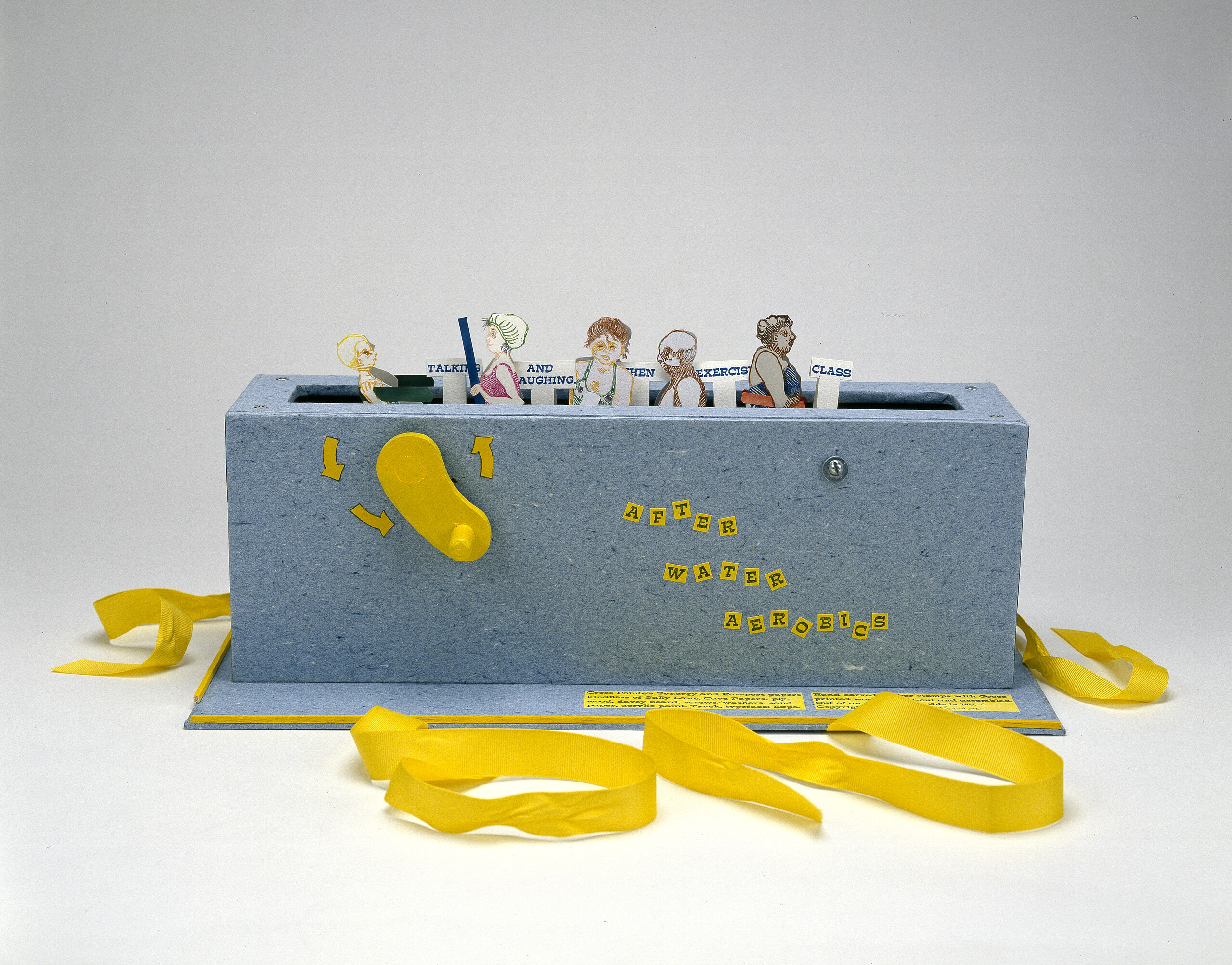 Paper figures in swimwear emerge from a grey-blue box with a slot in the top, like a toaster, and a yellow crank on the side. Small signs say “talking” “and laughing” “exercise” “class.” Yellow ribbons, untied, are at the base of the box.