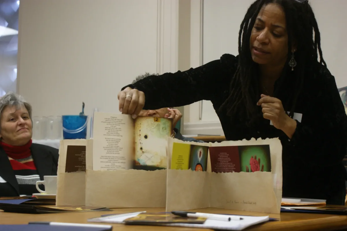 A dark-skinned woman with black dreadlocks and wearing a black sweater leans over a table and holds up a print of an artwork accompanied by a poem. To the left, a seated light-skinned woman with gray hair looks on.