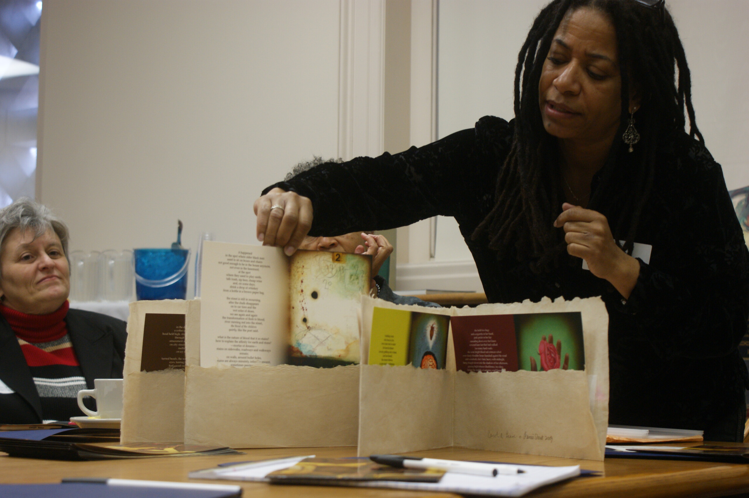 A dark-skinned woman with black dreadlocks and wearing a black sweater leans over a table and holds up a print of an artwork accompanied by a poem. To the left, a seated light-skinned woman with gray hair looks on.