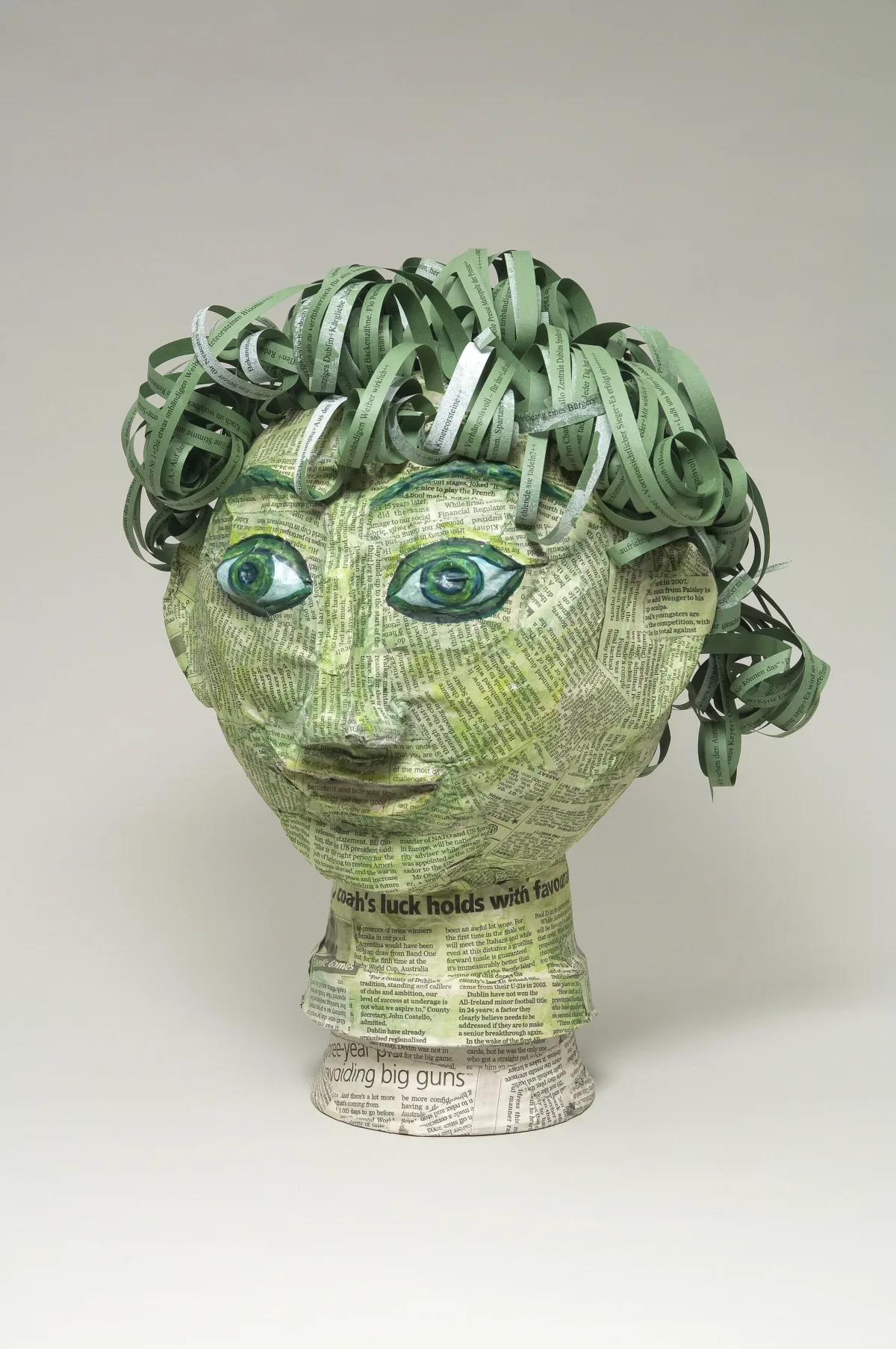 Papier-mâché head covered in clippings of pages from James Joyce's novel "Ulysses." In varying shades of green, the head includes curly, lettered strips for hair and wide and classic-Greek-style eyes under arched eyebrows.