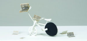 An all-white bicycle with a flat black sphere in the place of the rear wheel. A book is perched on a long arm extending from the handlebar mount, and pages of the book rest on the handlebars and are scattered on the ground.