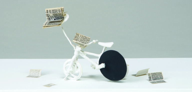 An all-white bicycle with a flat black sphere in the place of the rear wheel. A book is perched on a long arm extending from the handlebar mount, and pages of the book rest on the handlebars and are scattered on the ground.