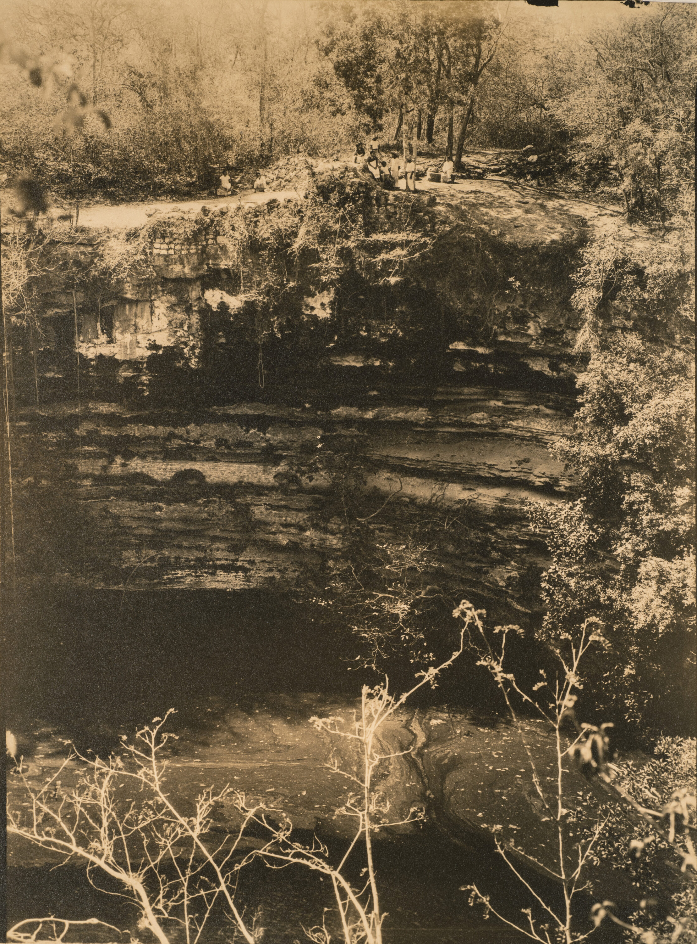 A sepia-toned vintage photograph of a pool of water below a vertical surface of natural rock. A group of figures dressed in white stand and sit on the flat space of ground at the top of the rock face.