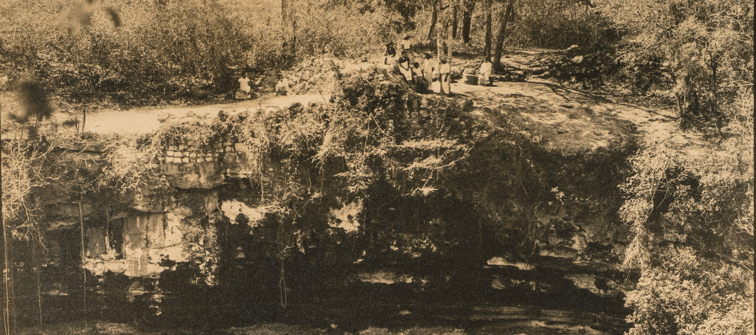 A sepia-toned vintage photograph of a pool of water below a vertical surface of natural rock. A group of figures dressed in white stand and sit on the flat space of ground at the top of the rock face.