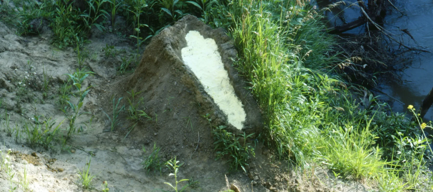 A mound of dirt in a grassy area by water. In the center of the mound is a human-shaped recess full of gunpowder.