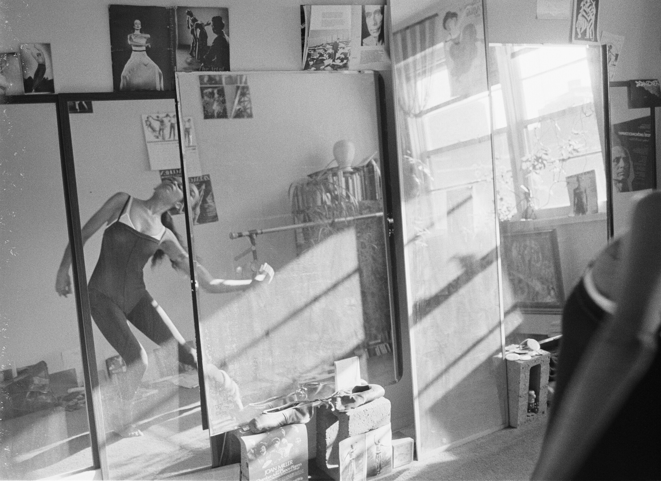 A black-and-white photograph of a woman dancing in a studio. The photo is taken behind the woman, facing a mirror, which shows the full scene of the studio and the woman's fluid move.