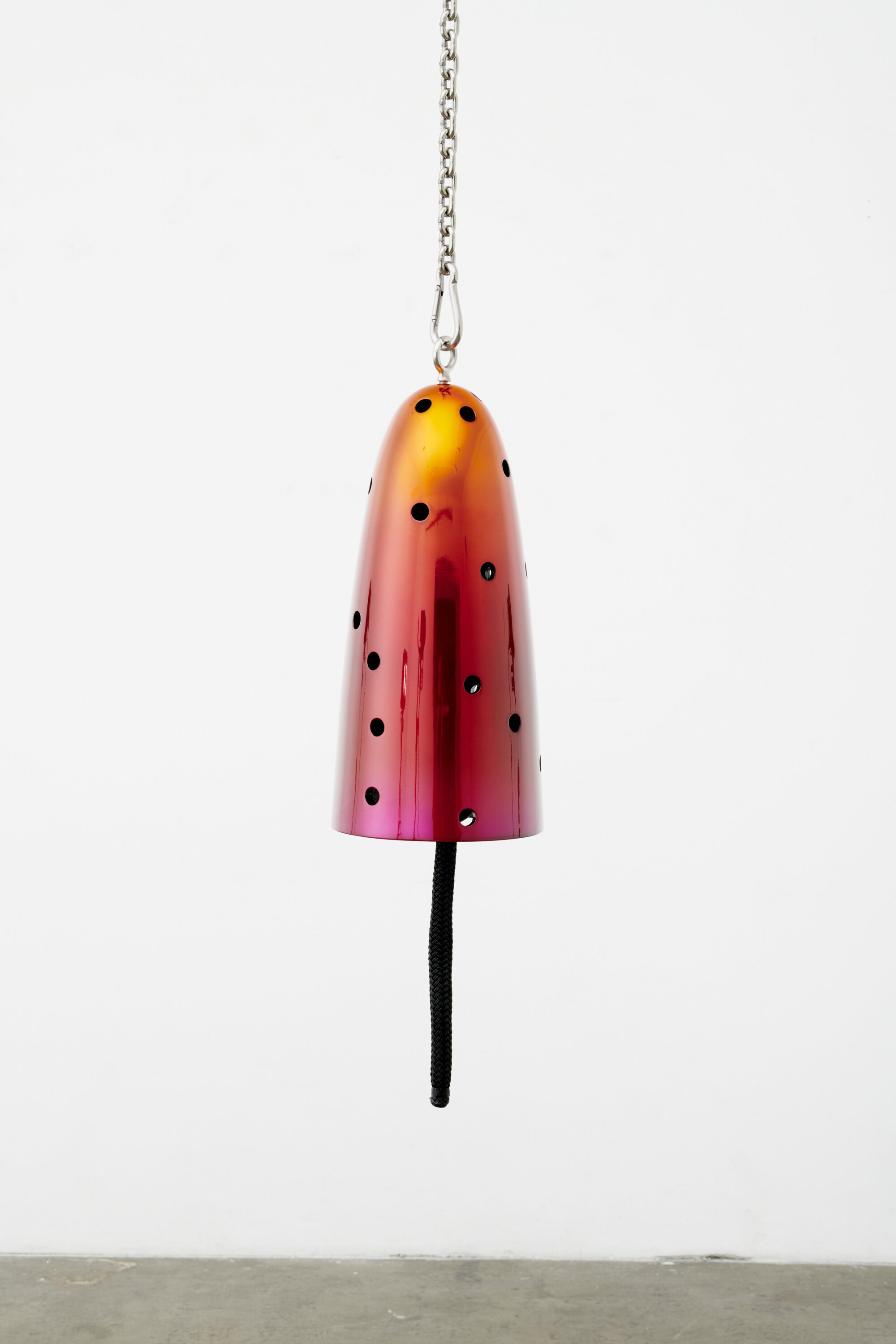 A bell-shaped sculpture in metallic shades of pink, red, and orange. The bell form is perforated and hangs from a silver metal chain.