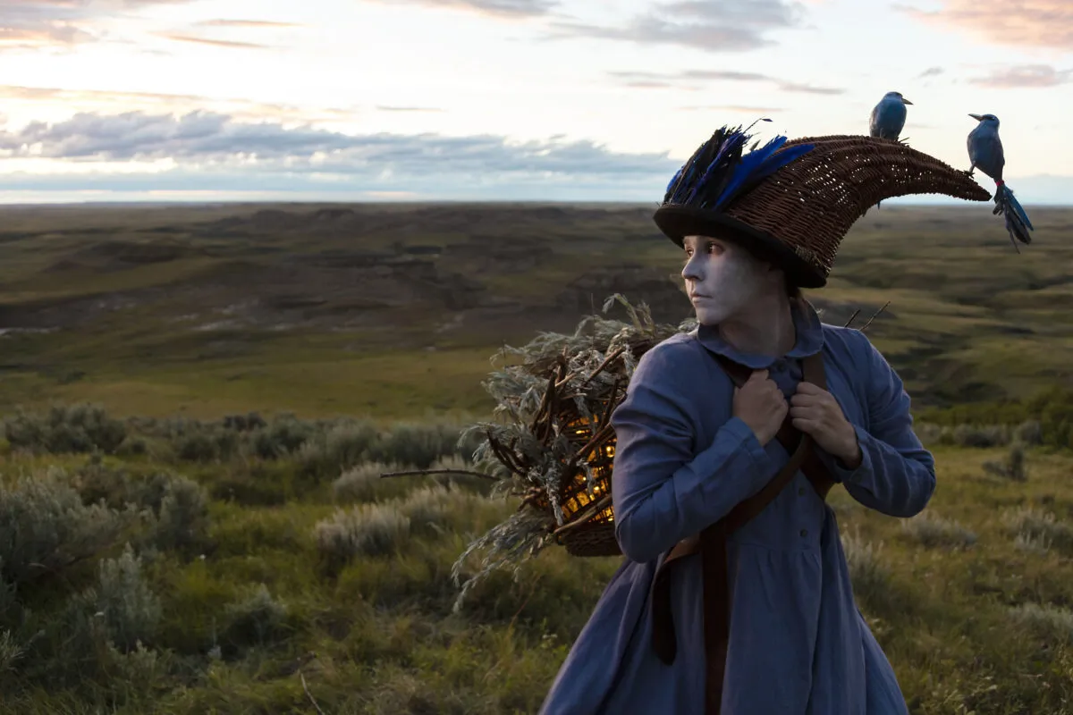 A woman in a blue dress and white face paint stands in a vast green landscape. She looks off to the side while carrying a foliage-covered lantern on her back. Two blue birds rest on her feather and wicker headdress.