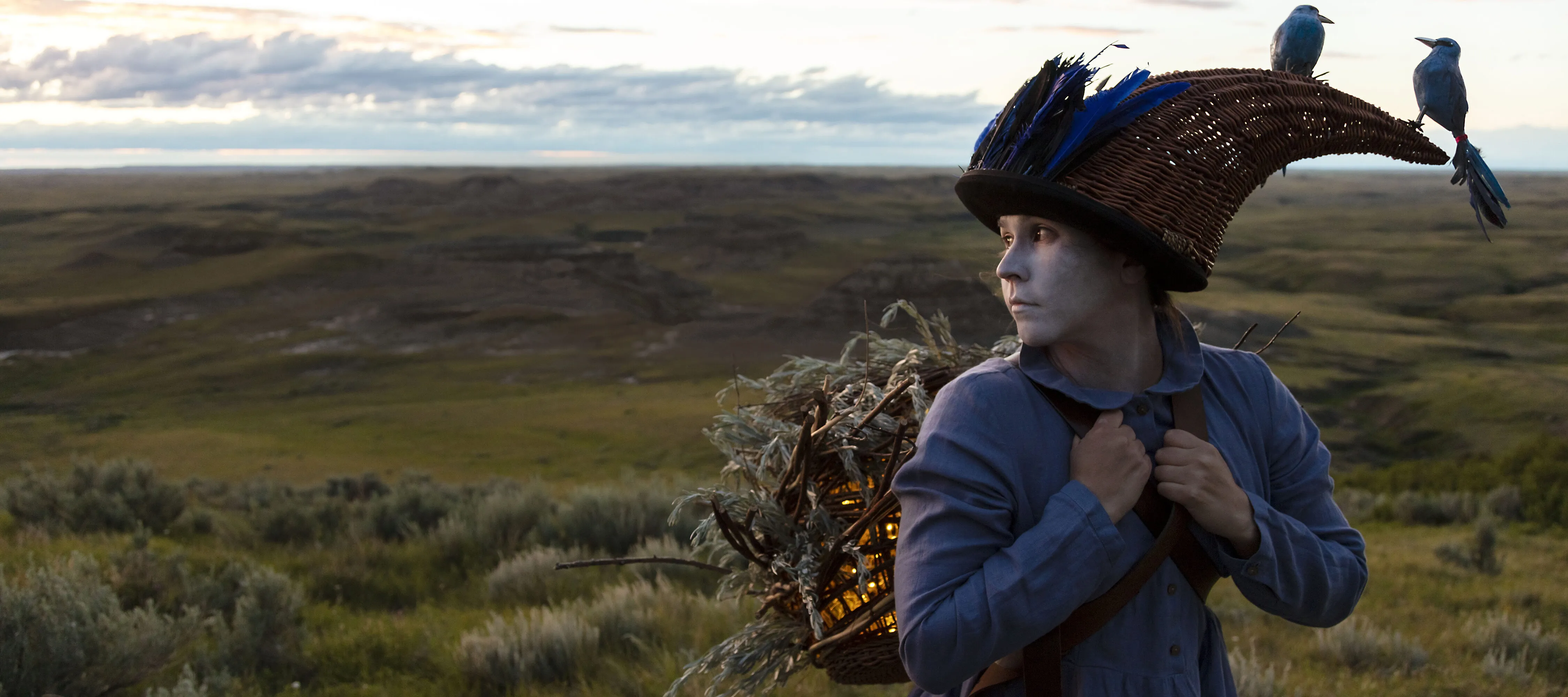 A woman in a blue dress and white face paint stands in a vast green landscape. She looks off to the side while carrying a foliage-covered lantern on her back. Two blue birds rest on her feather and wicker headdress.