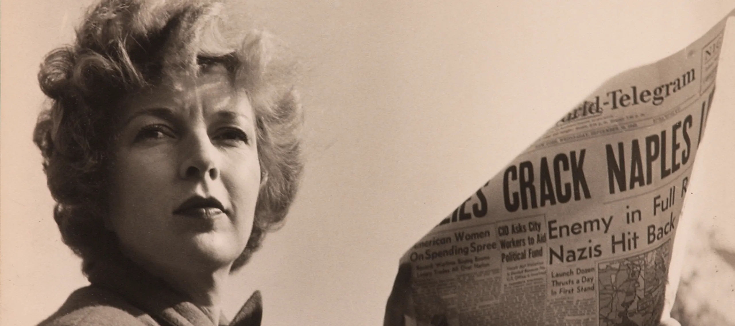 A black-and-white photograph of a light-skinned adult woman holding a newspaper with news about World War II. She wears a coat and her short, curly hair is caught in the wind.