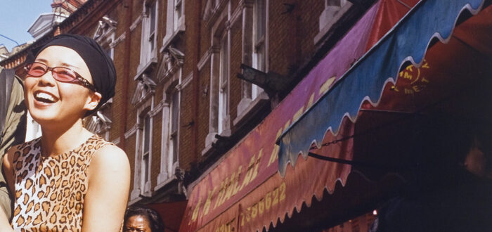 Color photograph of a street scene. Red brick buildings with colorful awnings are on the right and a smiling woman on the left. She holds the arm of a companion who has been cropped out of the left side of the image.