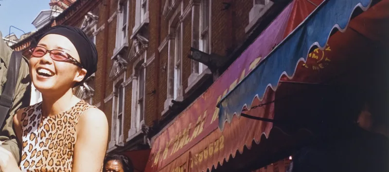 Color photograph of a street scene. Red brick buildings with colorful awnings are on the right and a smiling woman on the left. She holds the arm of a companion who has been cropped out of the left side of the image.